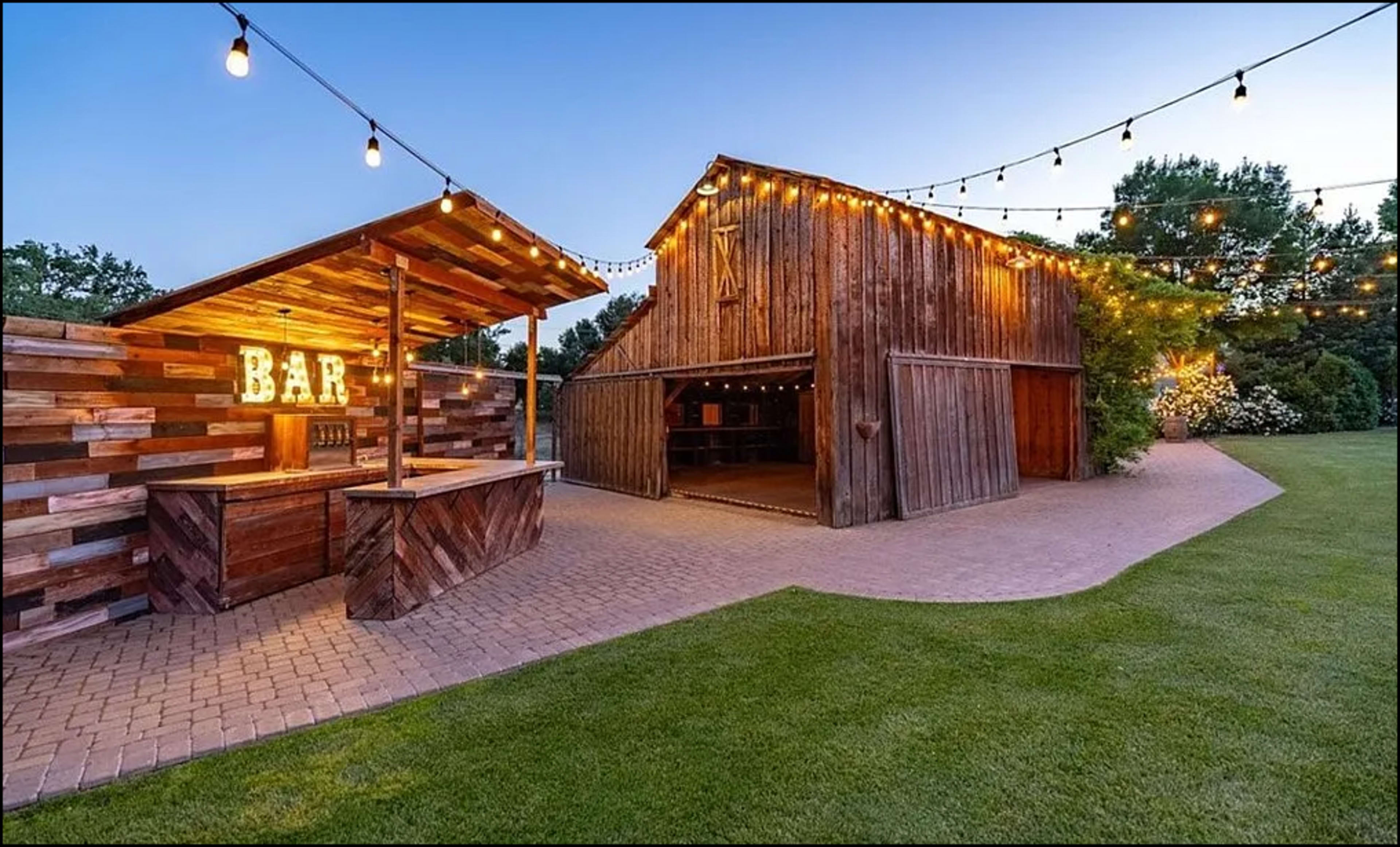 An outdoor bar made of wood with illuminated letters stands next to a rustic barn, surrounded by green grass and string lights in the evening.