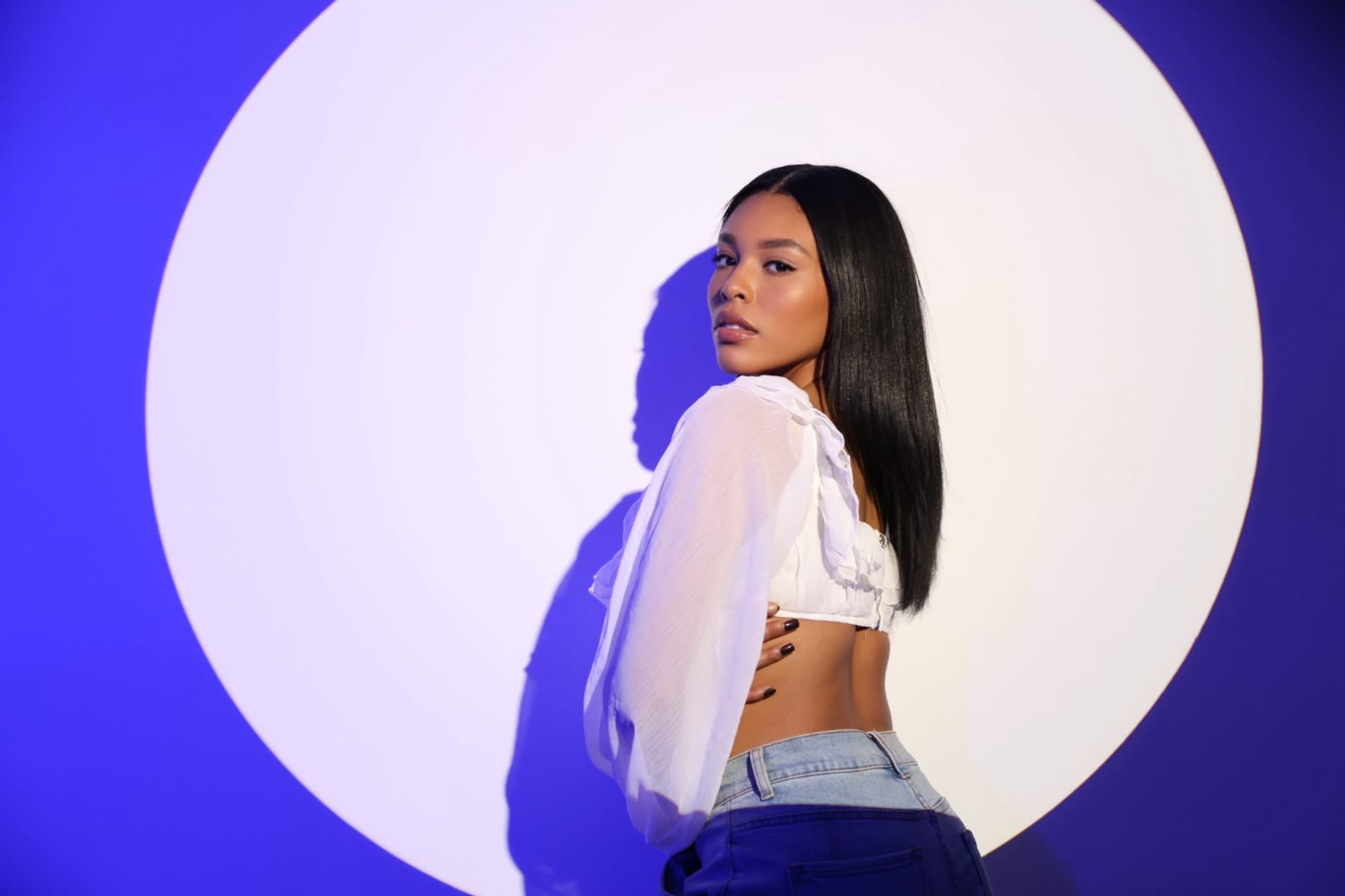 A person with long black hair poses in front of a large circular light creation, wearing a white top and blue jeans.