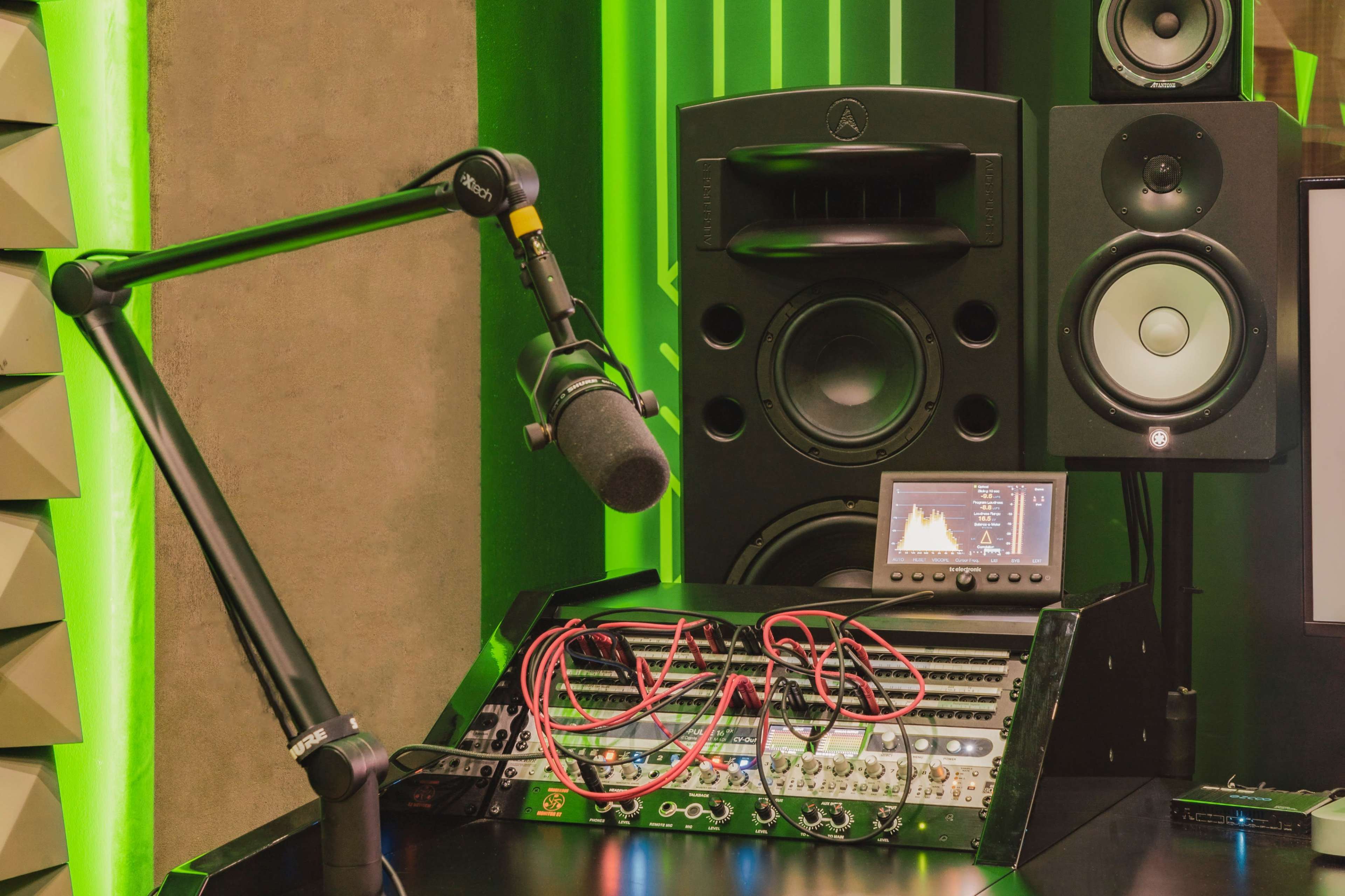 The image shows a recording studio setup featuring a microphone mounted on a boom arm, audio monitors, and a mixer with connected cables.