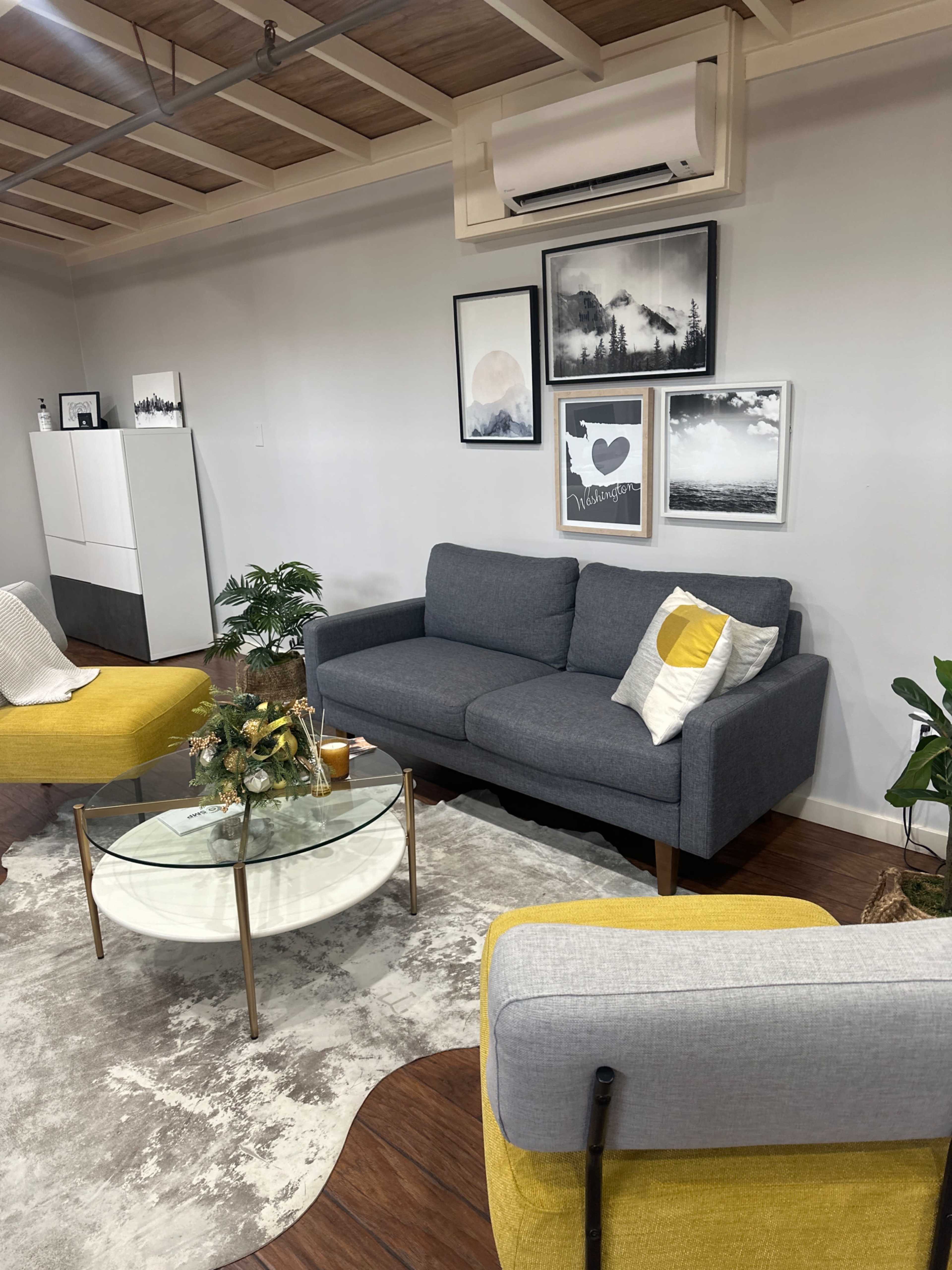 The image shows a modern living room with a gray sofa, yellow accent chairs, a glass coffee table, and framed artwork on the walls.