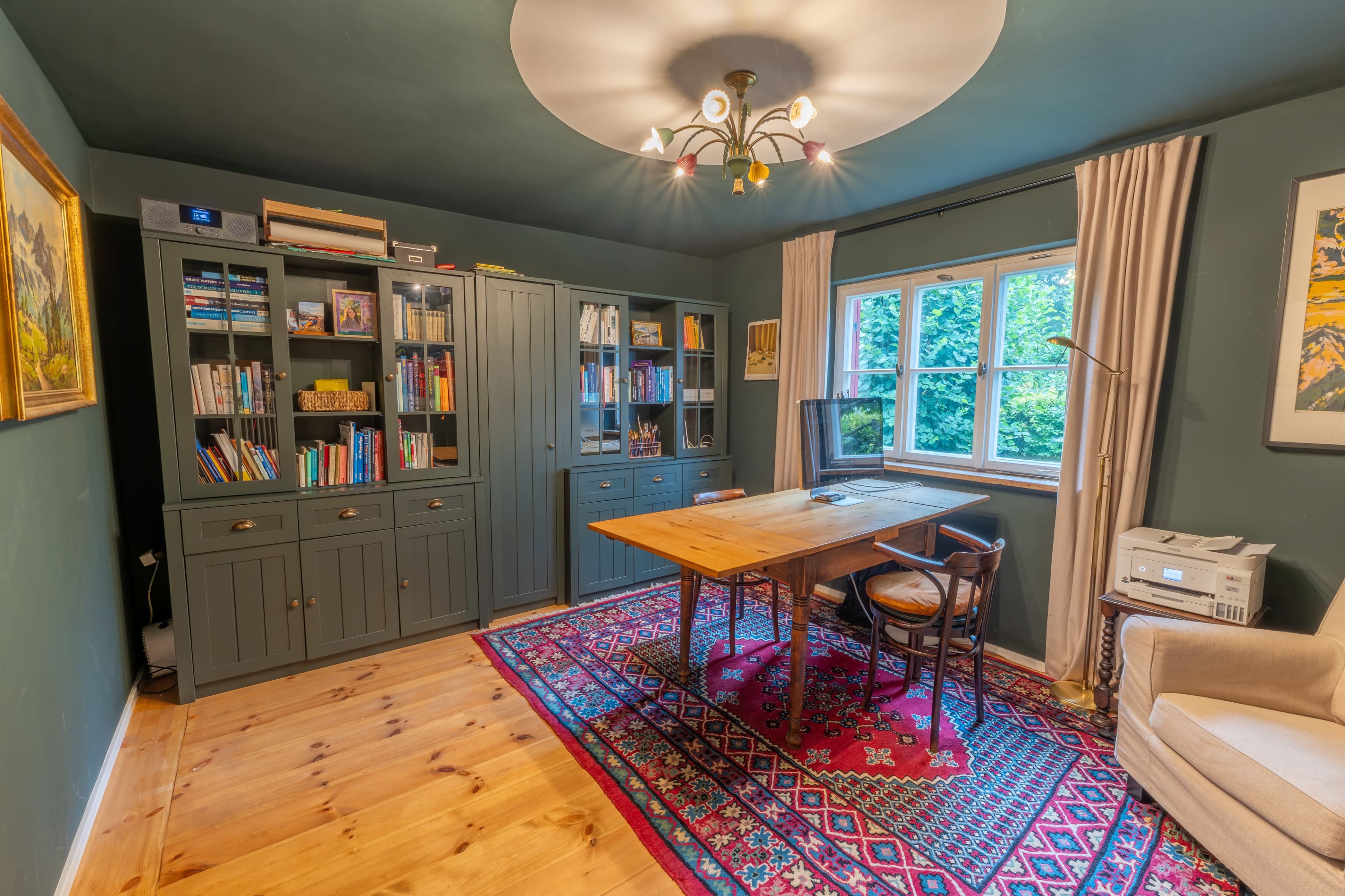 The image shows a cozy home office with a large wooden table, bookshelves filled with books, a computer setup, and a comfortable chair, all set against a green wall.