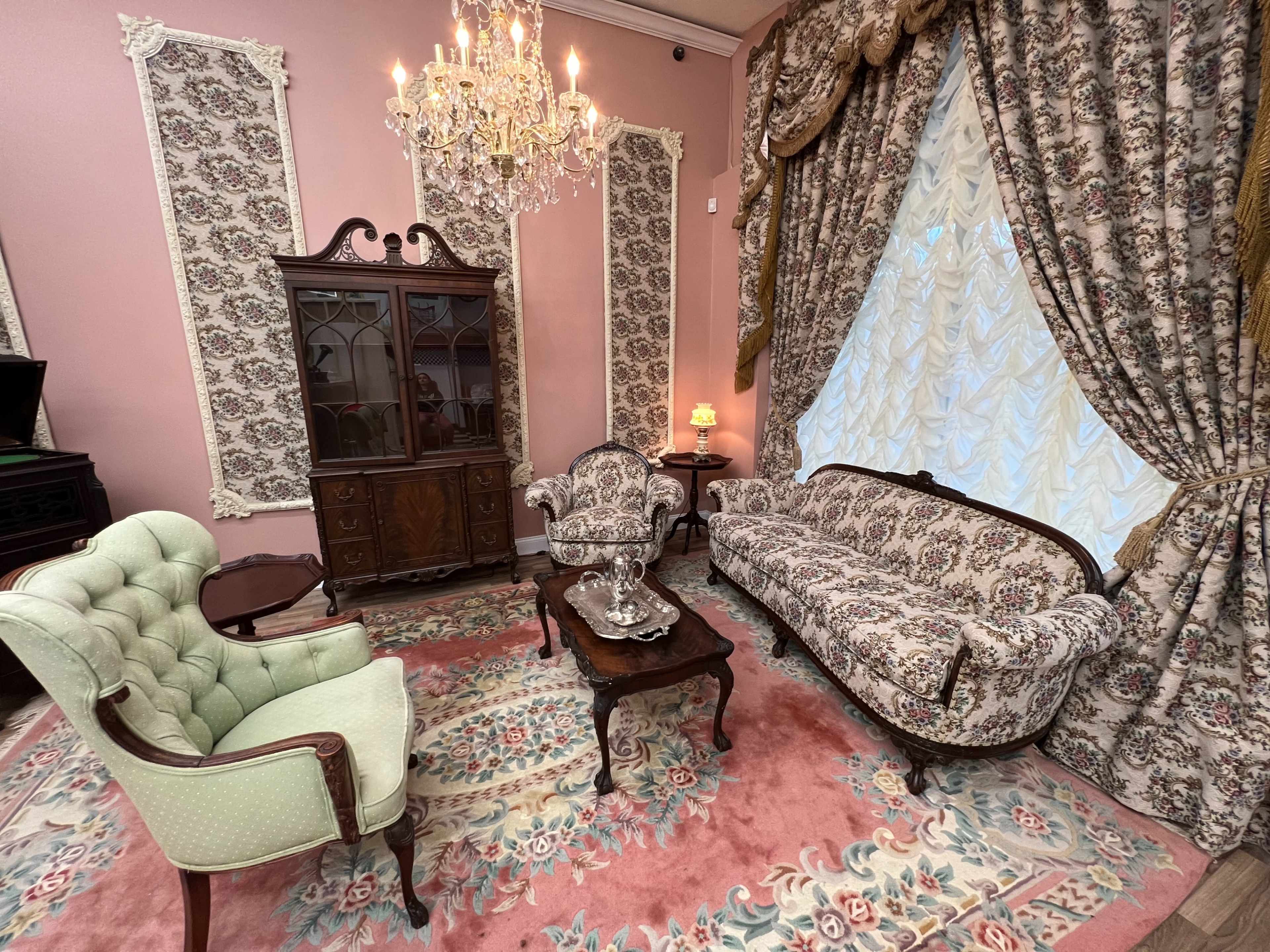 A vintage sitting room with ornate furniture, including a floral patterned sofa and armchairs, a dark wooden cabinet, and a chandelier, all set against pink walls and a patterned rug.