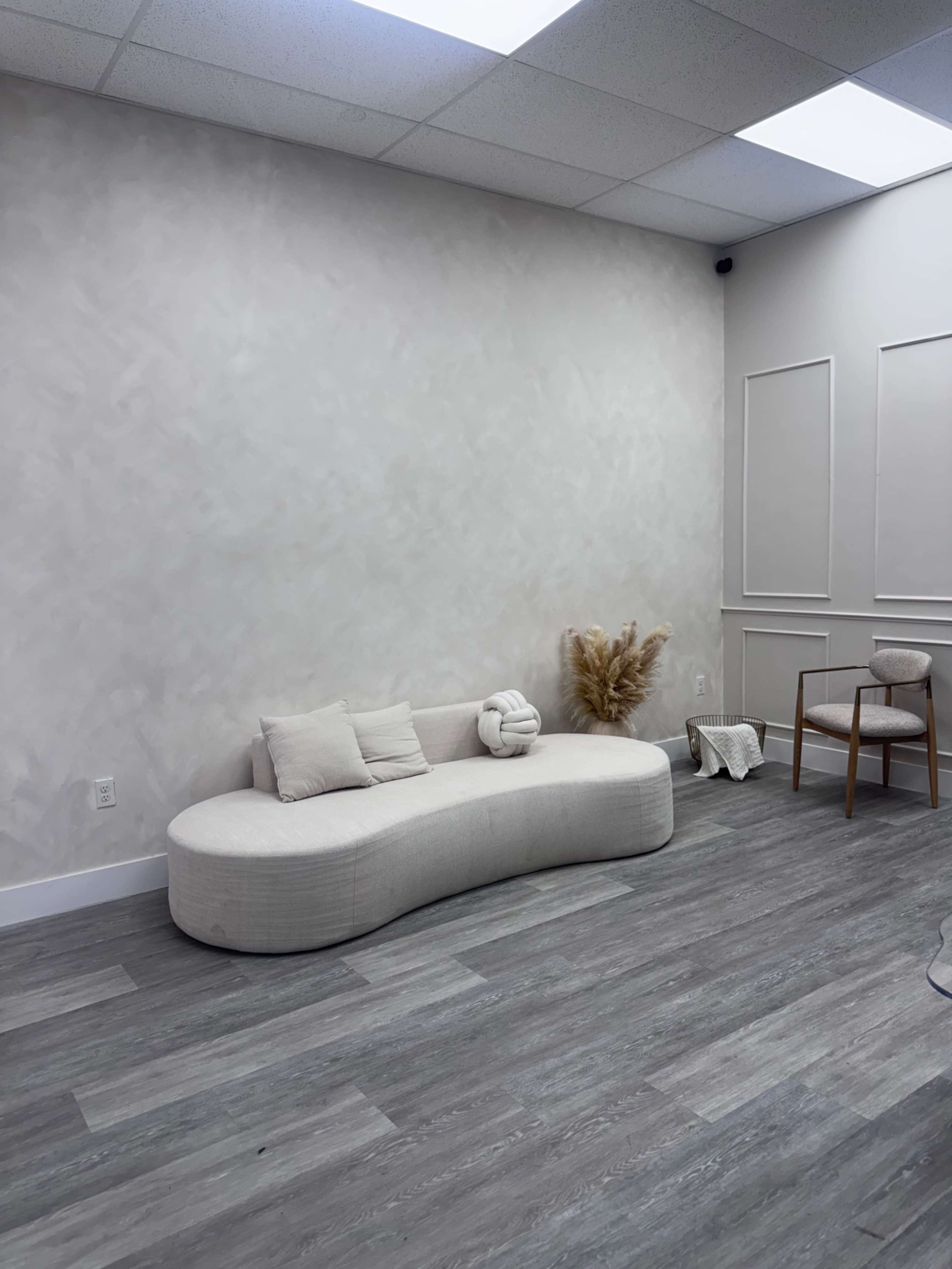 The image shows a minimalist room featuring a curved white couch, a wooden chair, and decorative pampas grass against a light-colored wall.