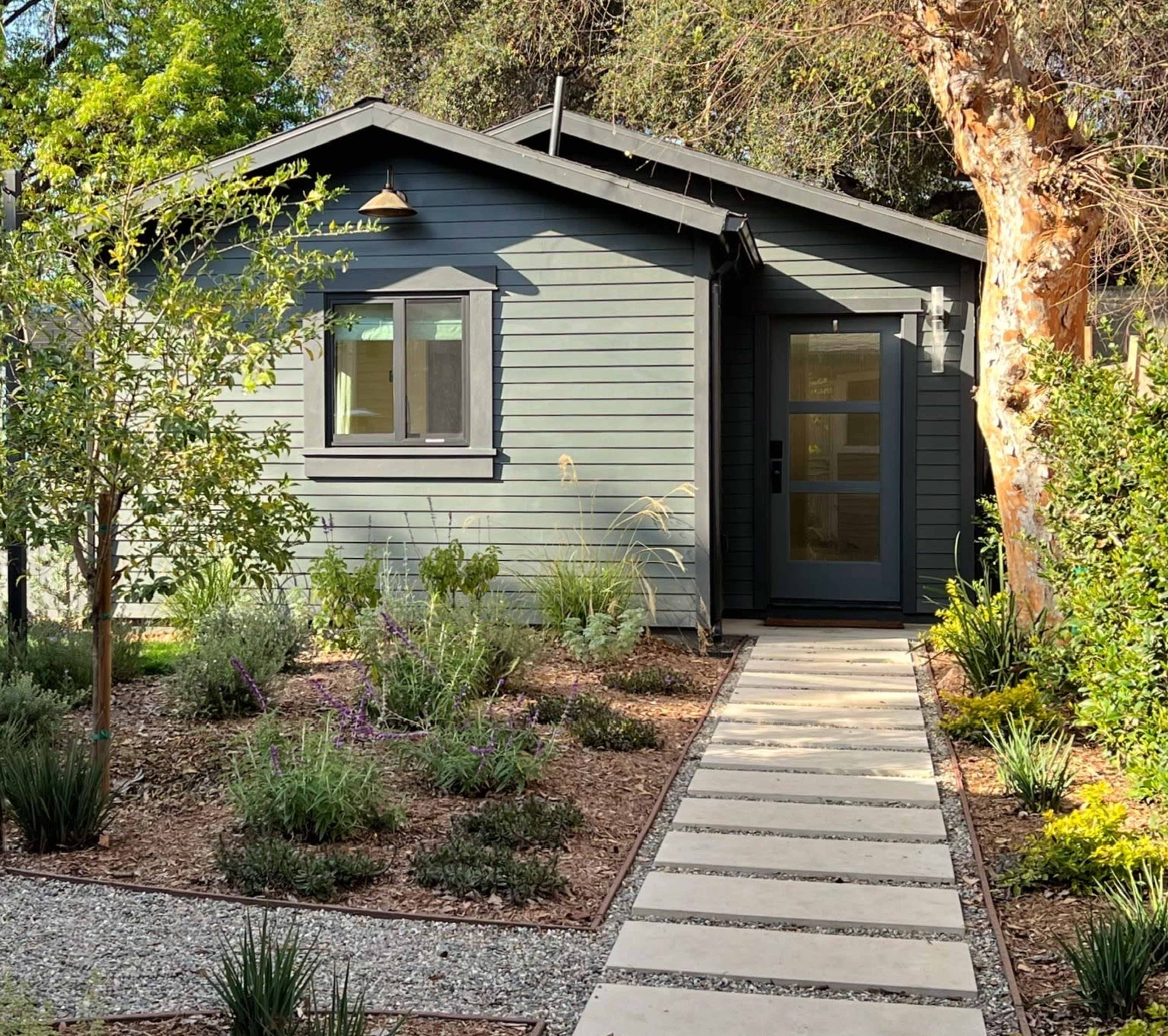 A gray-blue single-story house with a dark front door and a stone path is surrounded by lush landscaping and greenery.