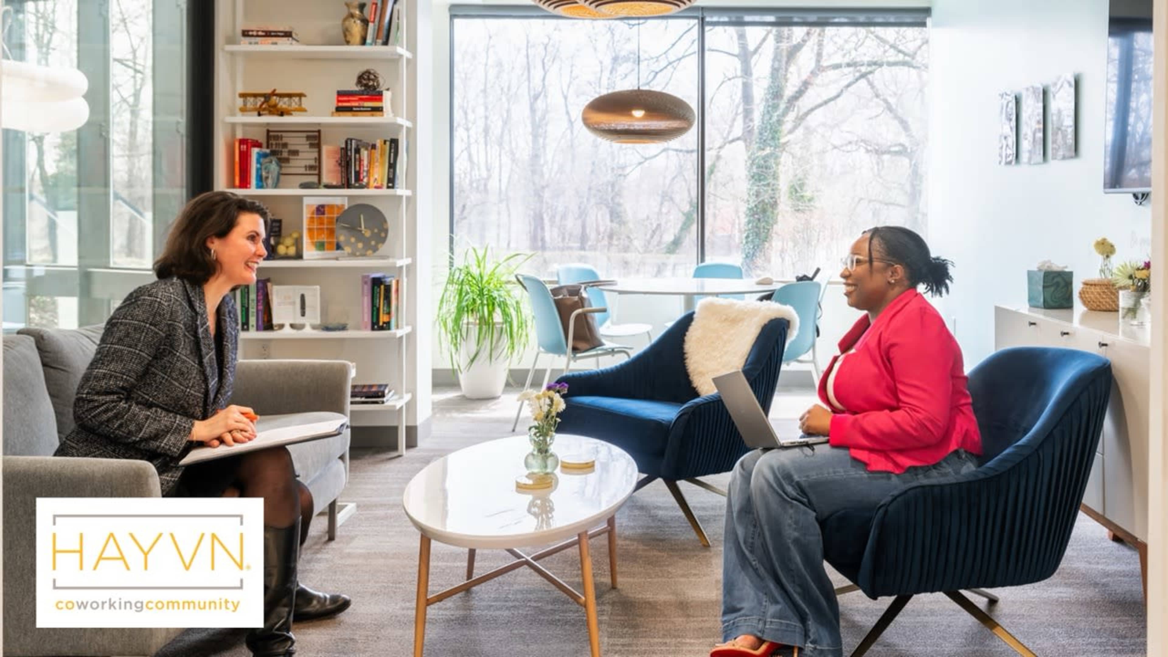 Two women are seated in a bright coworking space, engaging in a conversation while surrounded by bookshelves and large windows that overlook a natural setting.