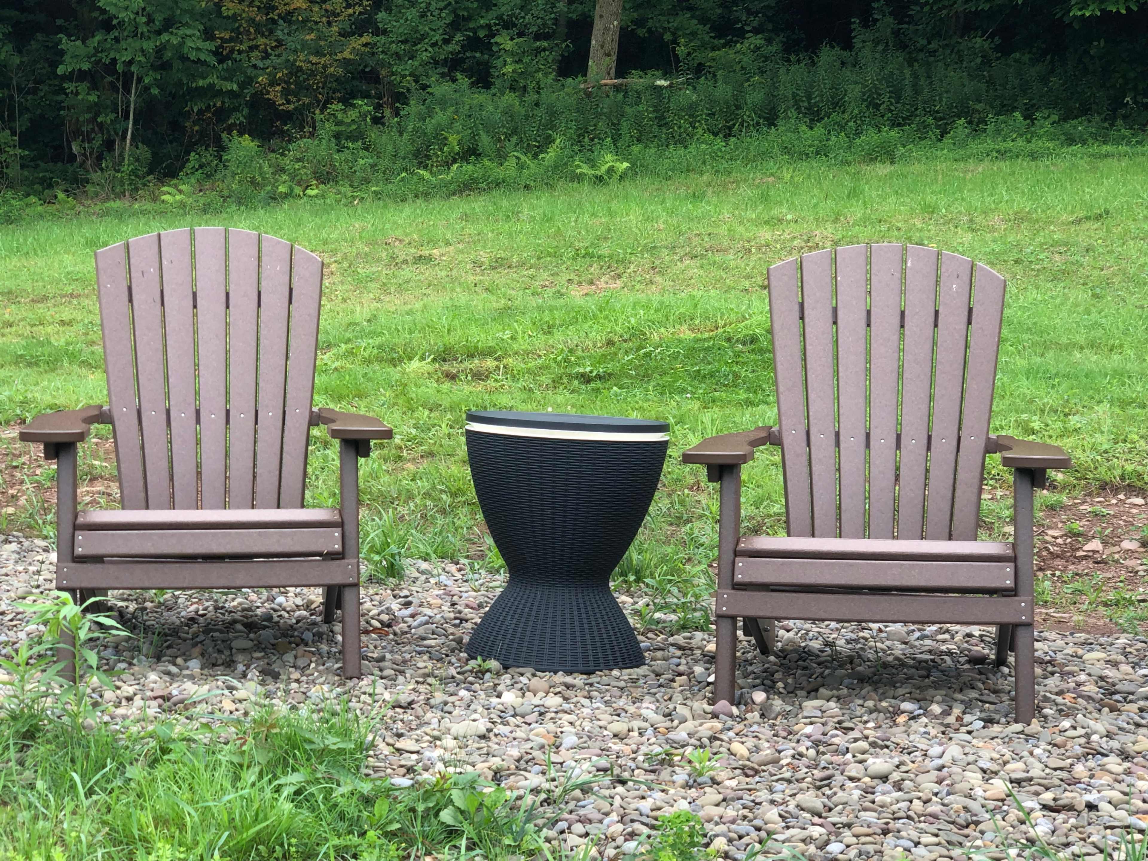 Two Adirondack chairs flank a round black side table on a gravel patch surrounded by a grassy area.