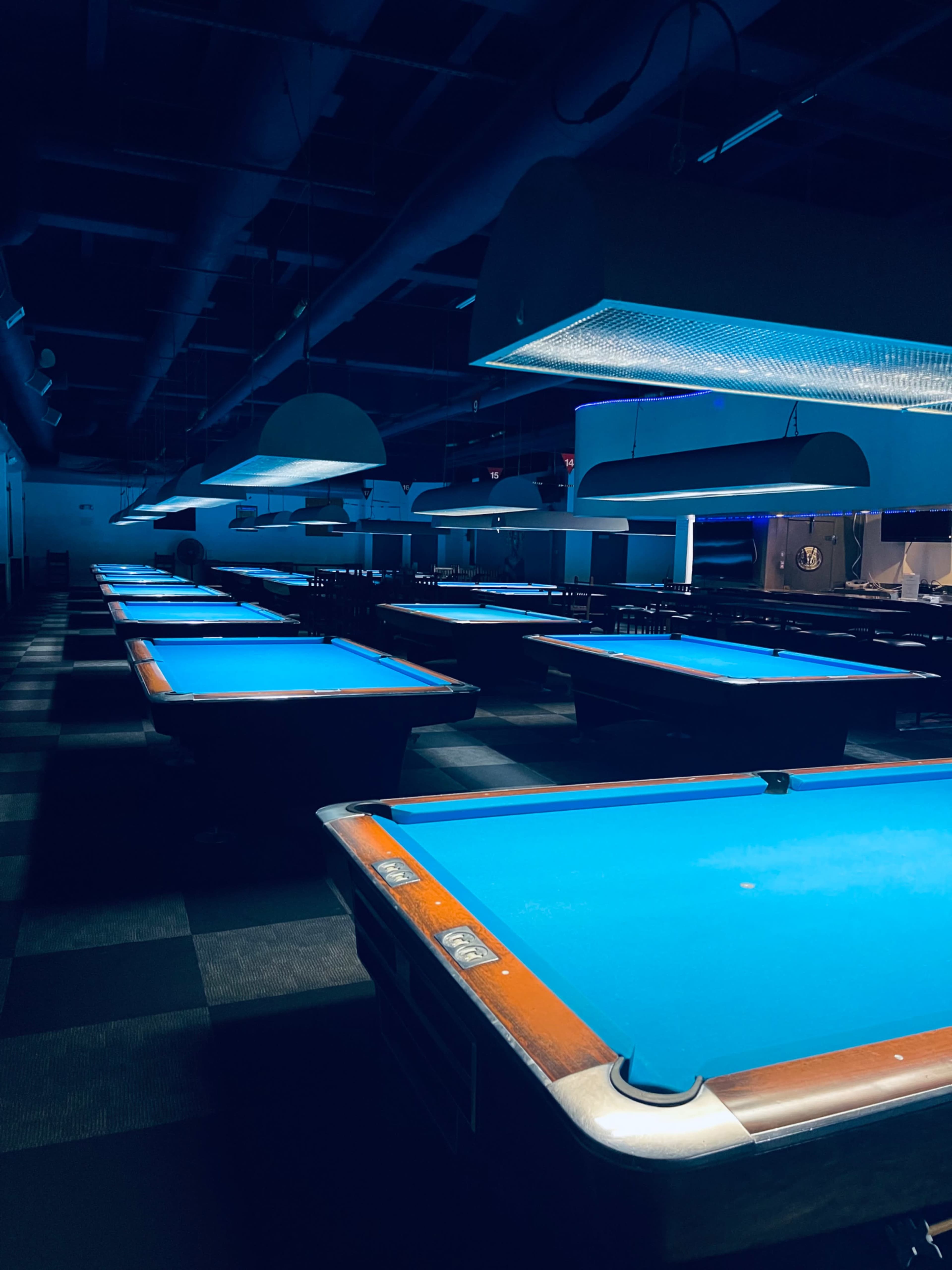 A dimly lit room filled with pool tables, each illuminated by overhead lights, casting a blue glow over the table surfaces.