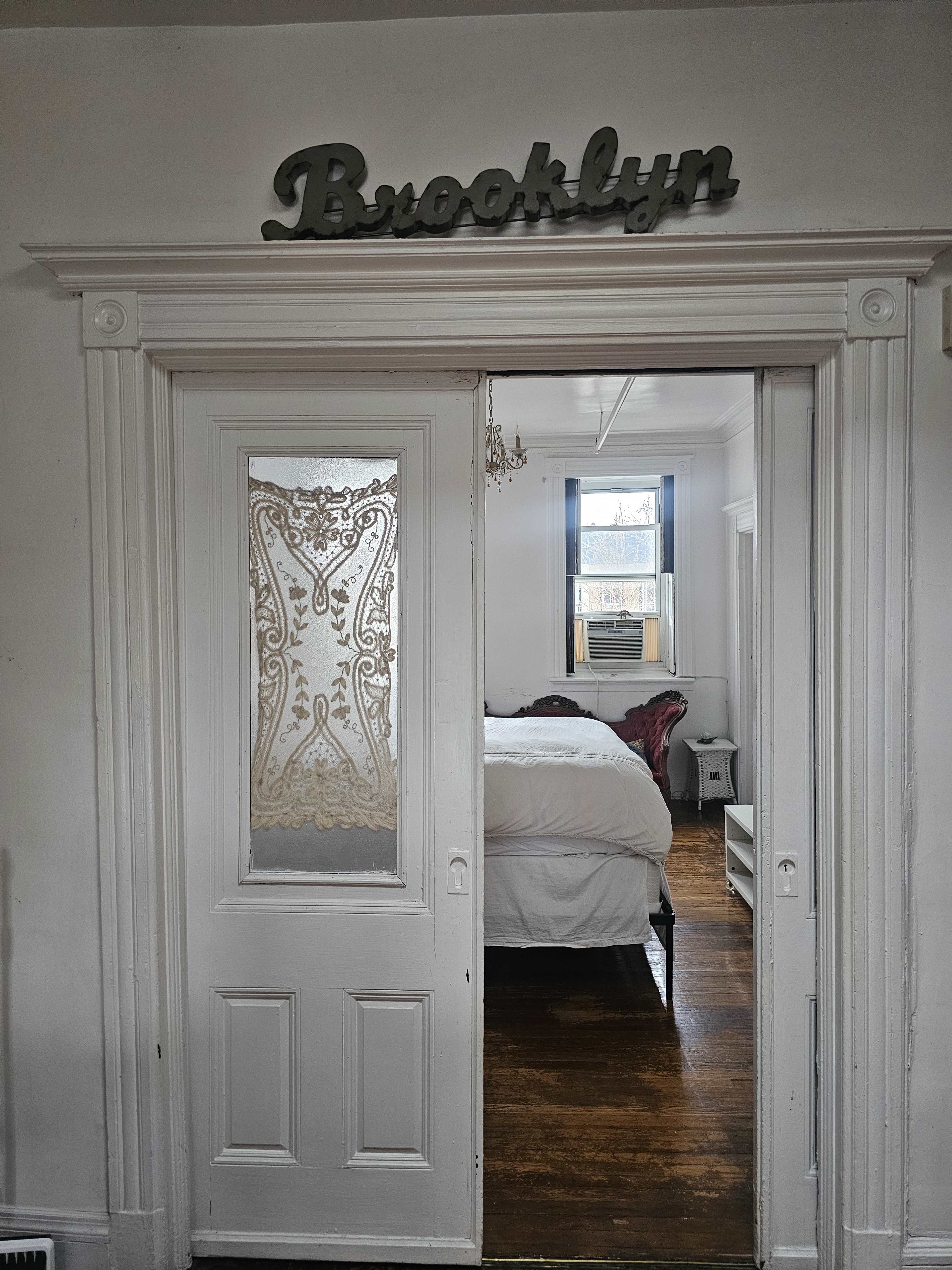 The image shows a white doorway with a decorative glass panel labeled "Brooklyn," leading into a bedroom with a white bed and a vintage chair.