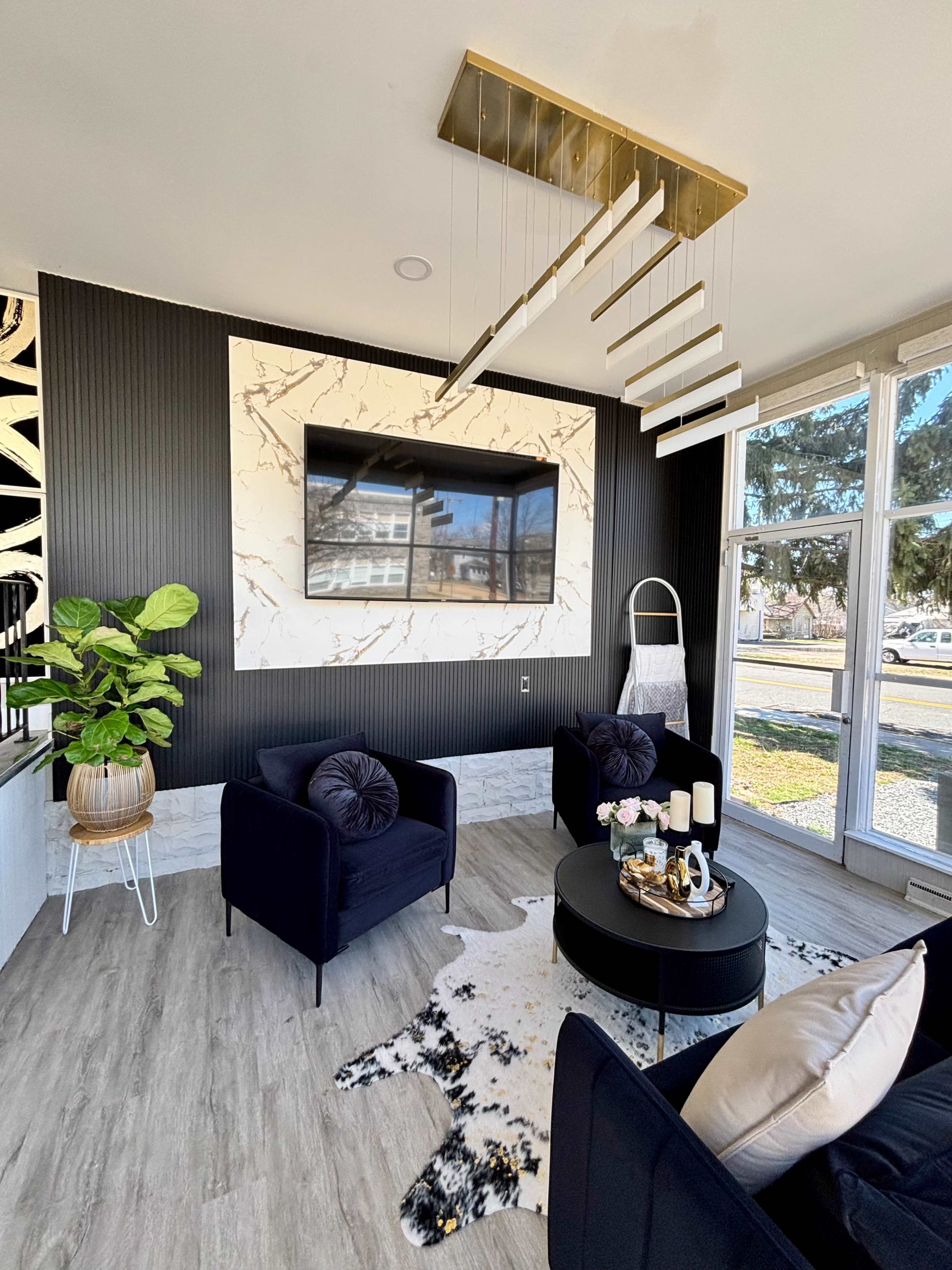 The image shows a modern living room with a black and white color scheme, featuring a large wall-mounted TV above a marble panel, black chairs, and a circular coffee table.