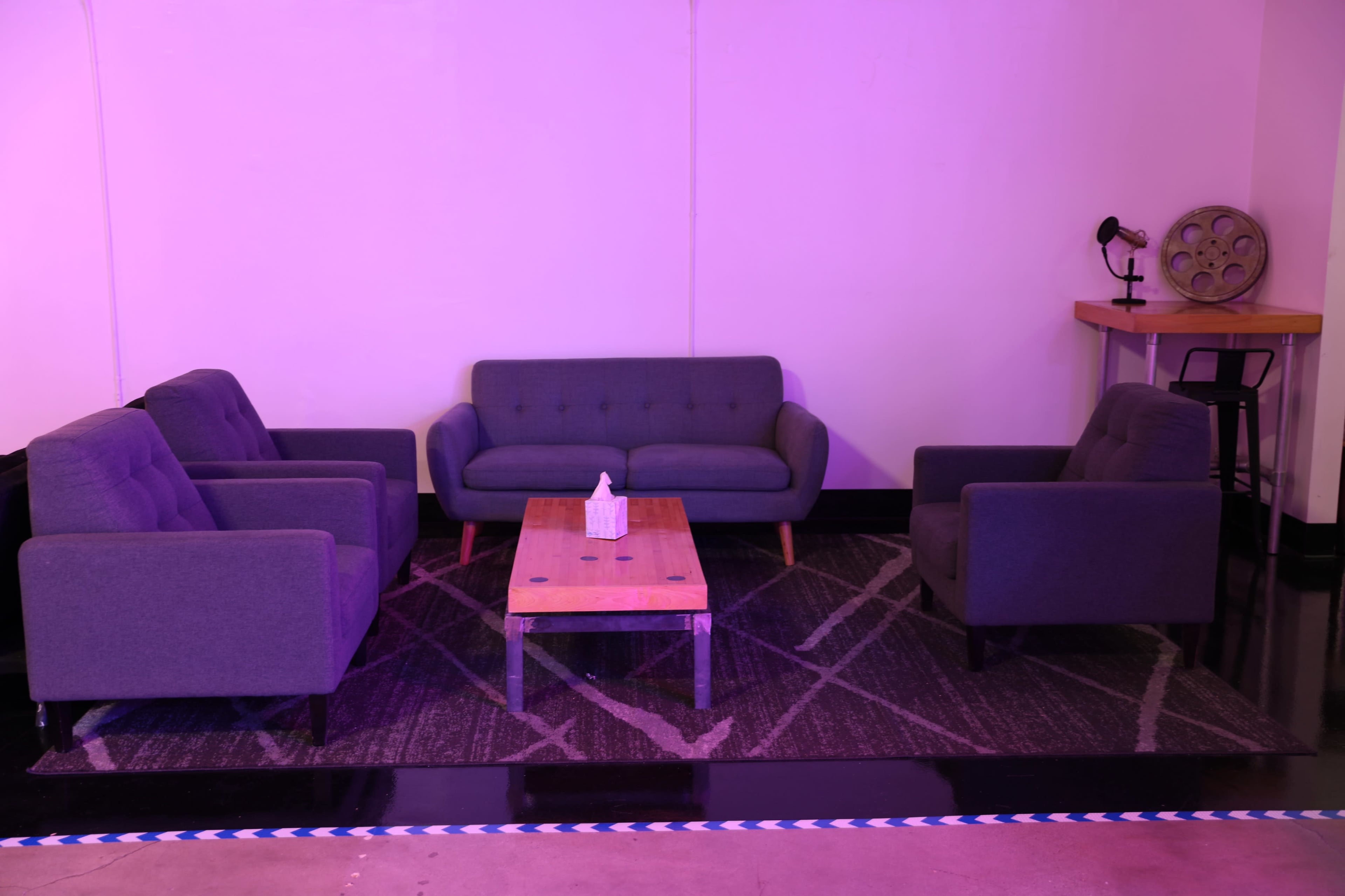 A modern seating area features a gray sofa and four matching chairs arranged around a wooden coffee table on a patterned rug, illuminated by soft purple light.