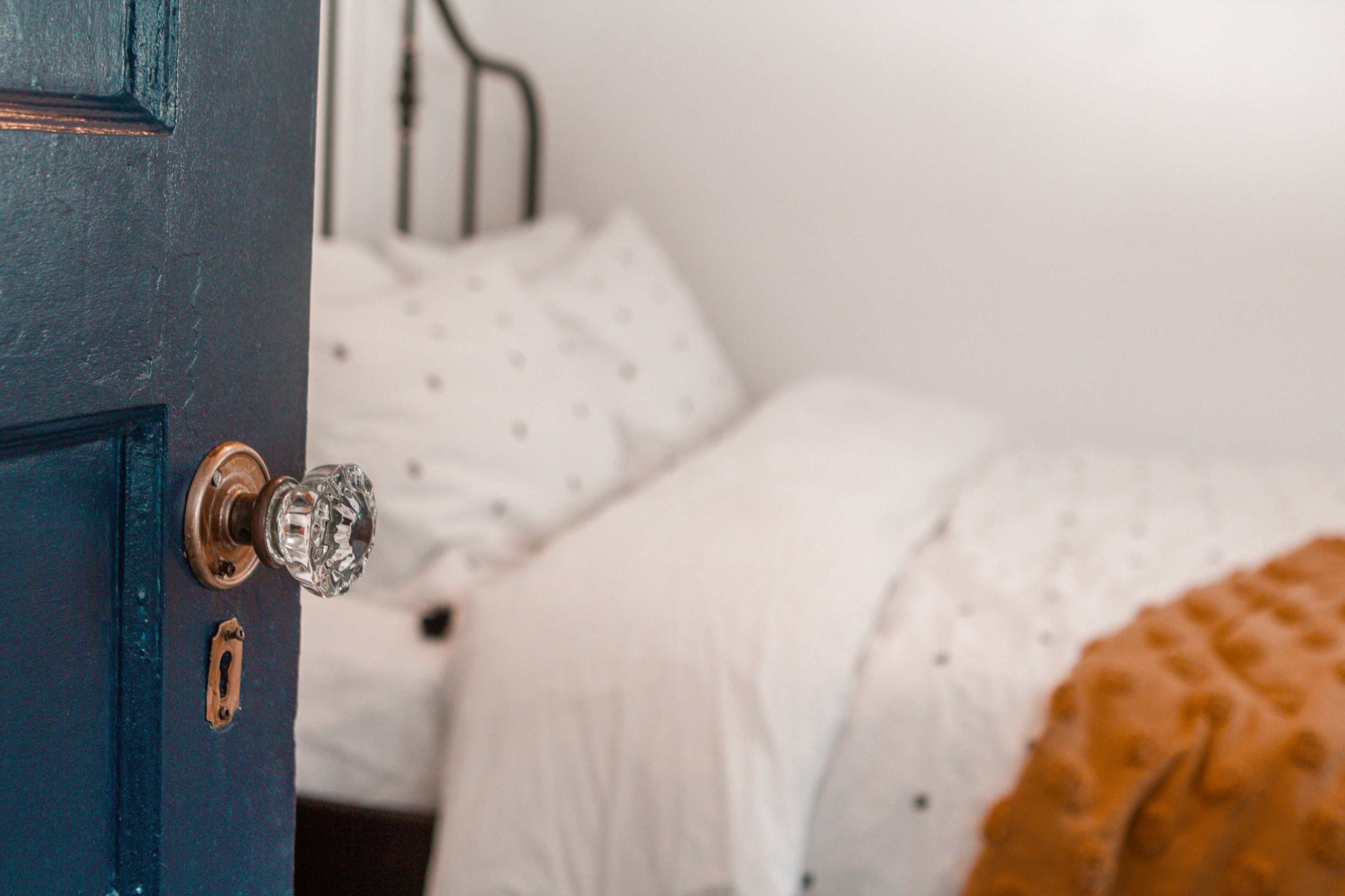A blue door is partially open, revealing a bed with white bedding and a brown throw blanket in a softly lit room.