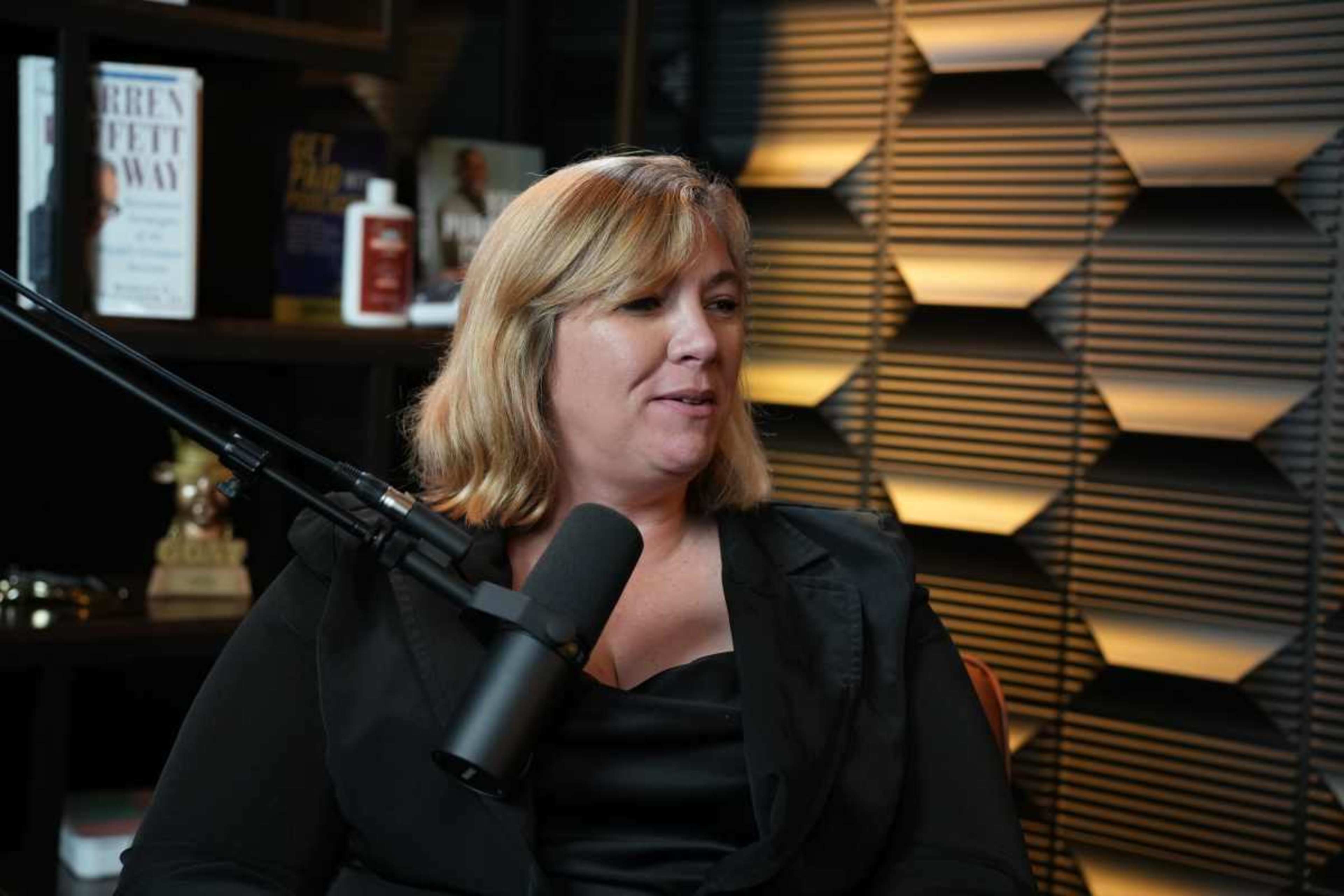 A woman is speaking into a microphone in a well-lit studio, surrounded by books and decorative items on shelves.