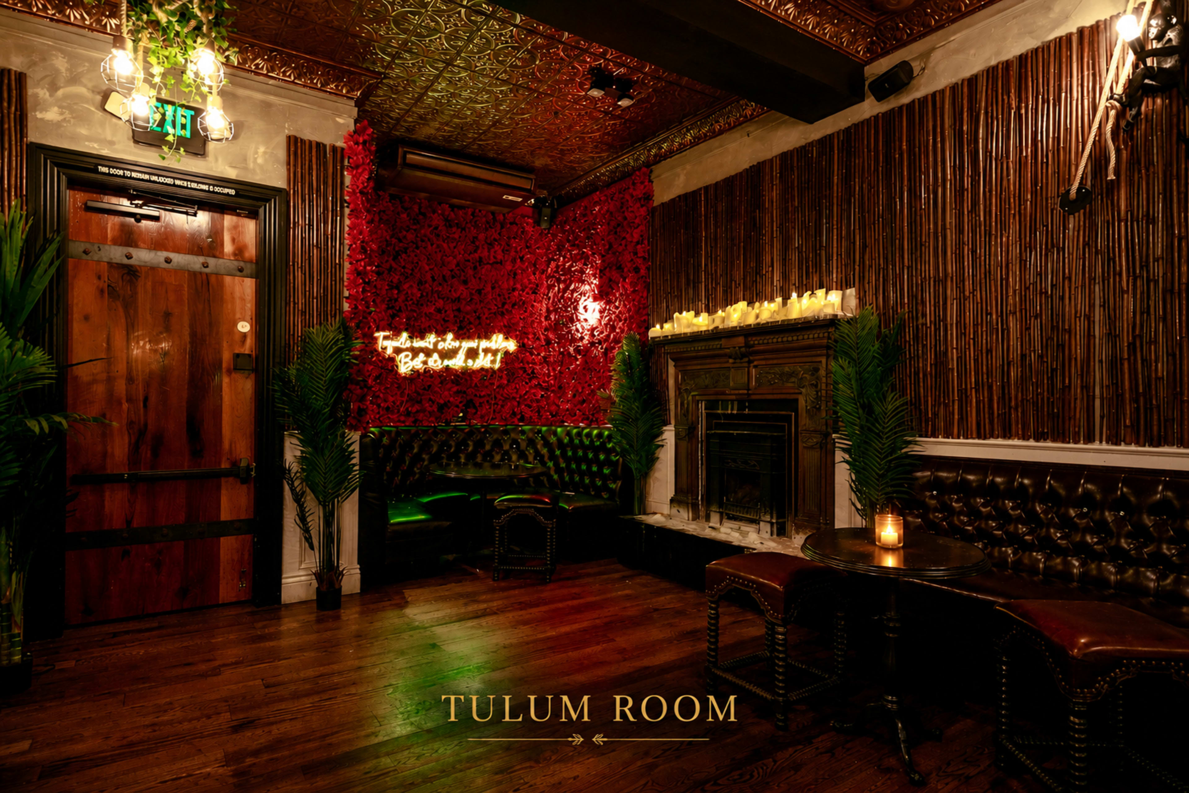 The image depicts an elegantly decorated bar room named "Tulum Room," featuring a red floral wall, dark wood accents, and a seating area with lush greenery and soft lighting.