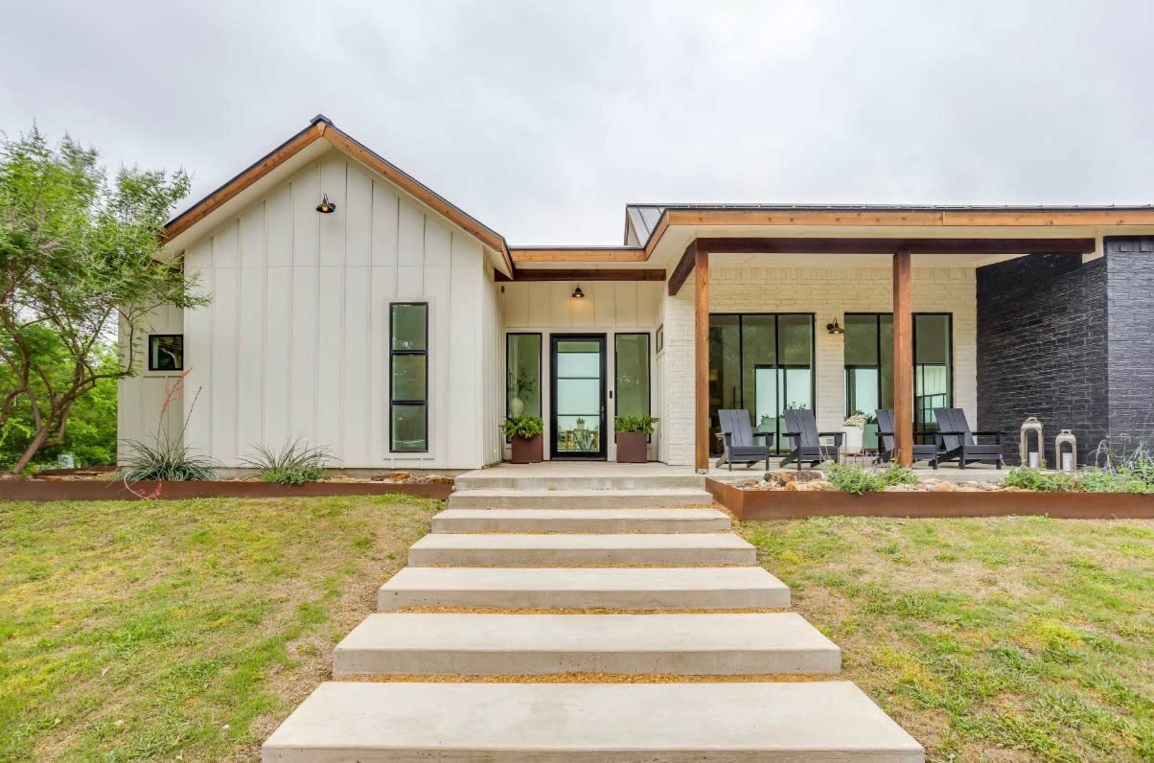 The image shows a modern house with a combination of white and black exterior walls, featuring a pathway leading to an entrance framed by large windows and a porch with chairs.
