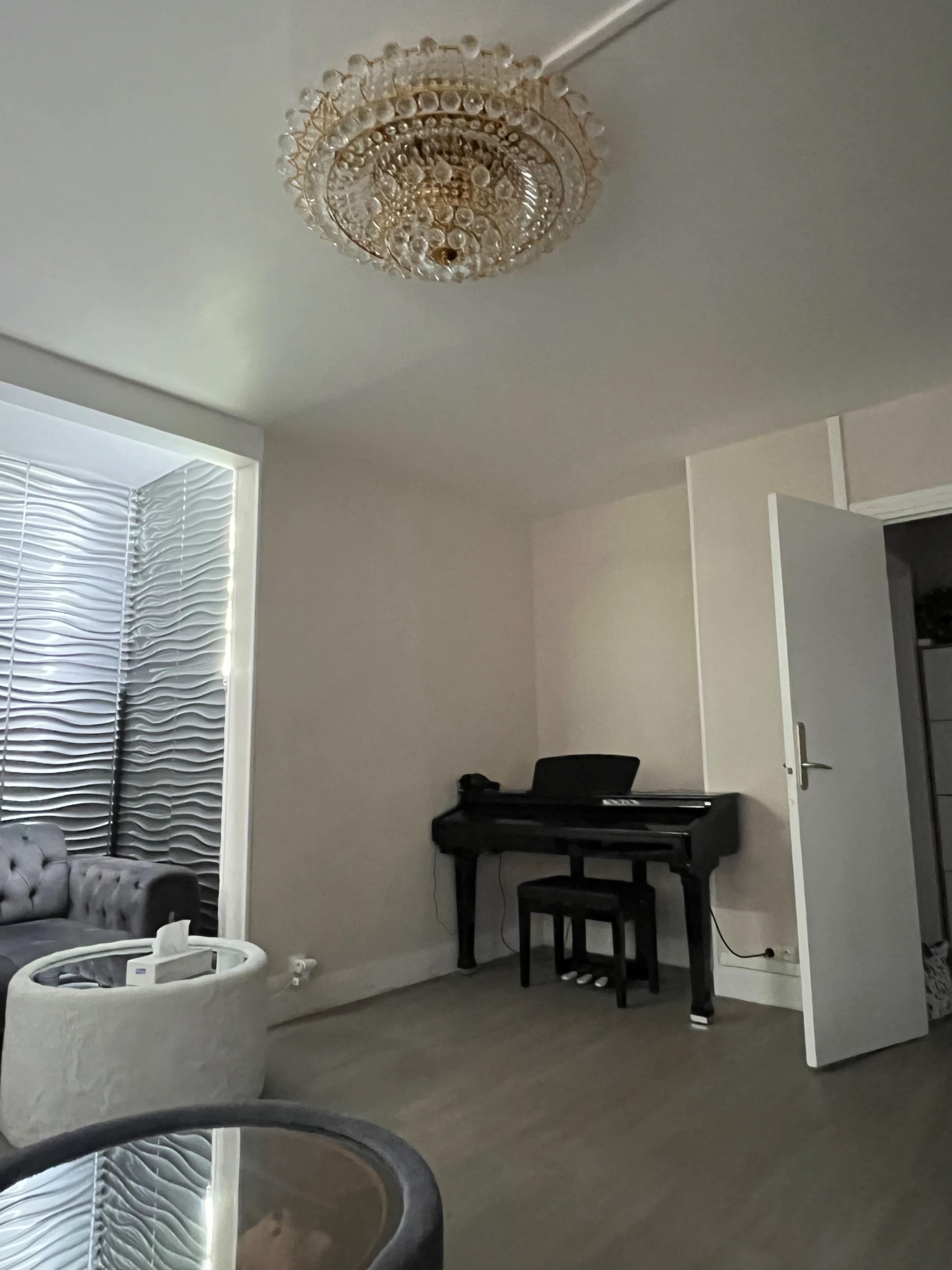 The image shows a minimalist living room featuring a chandelier, a piano, and modern furniture.