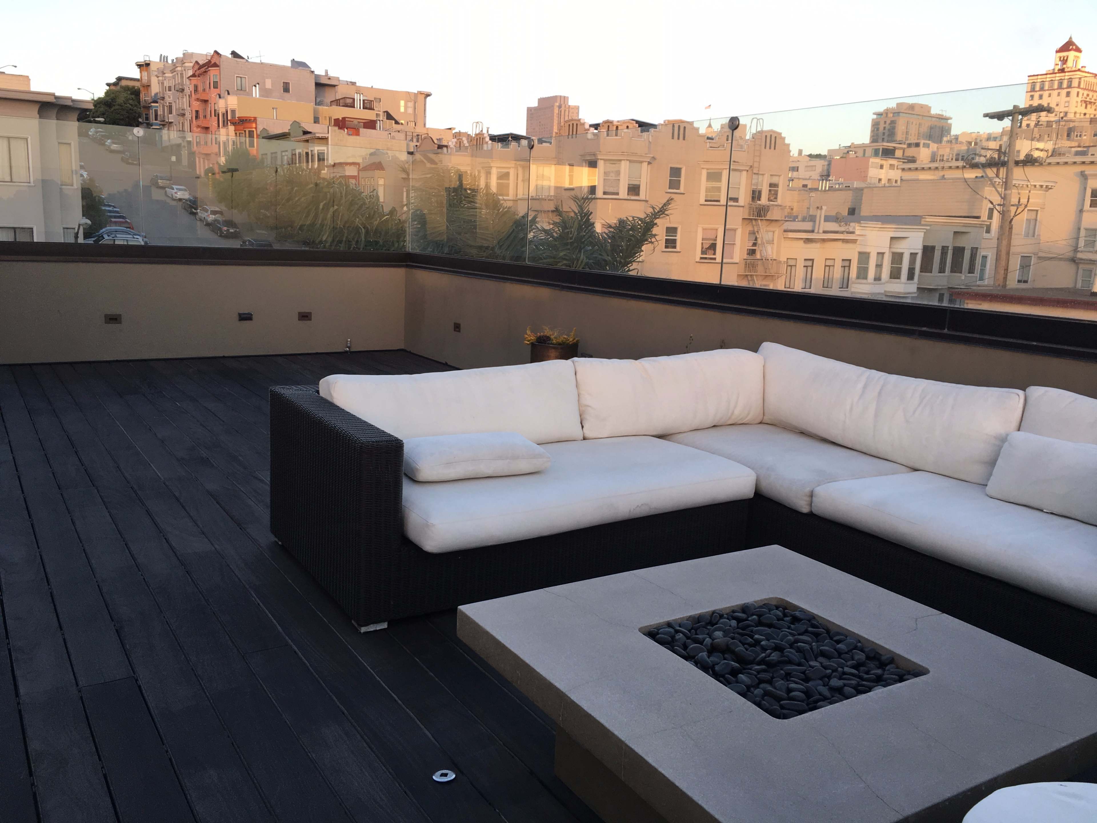 A rooftop terrace features a white sectional sofa and a fire pit surrounded by small black stones, with city buildings visible in the background.