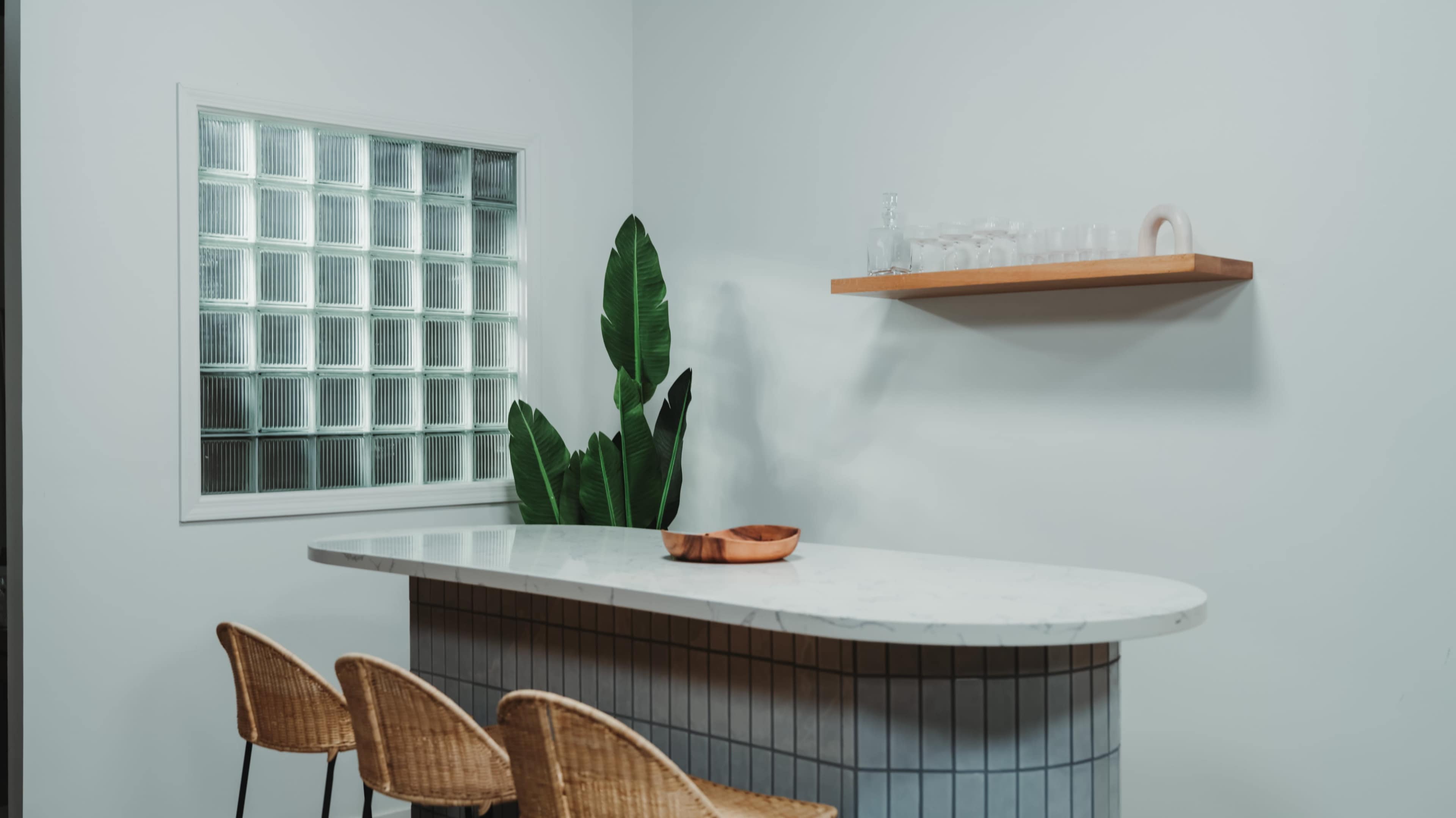 A modern kitchen features a marble countertop with three wicker chairs, a glass block window, and a wooden shelf displaying glassware and a bowl.