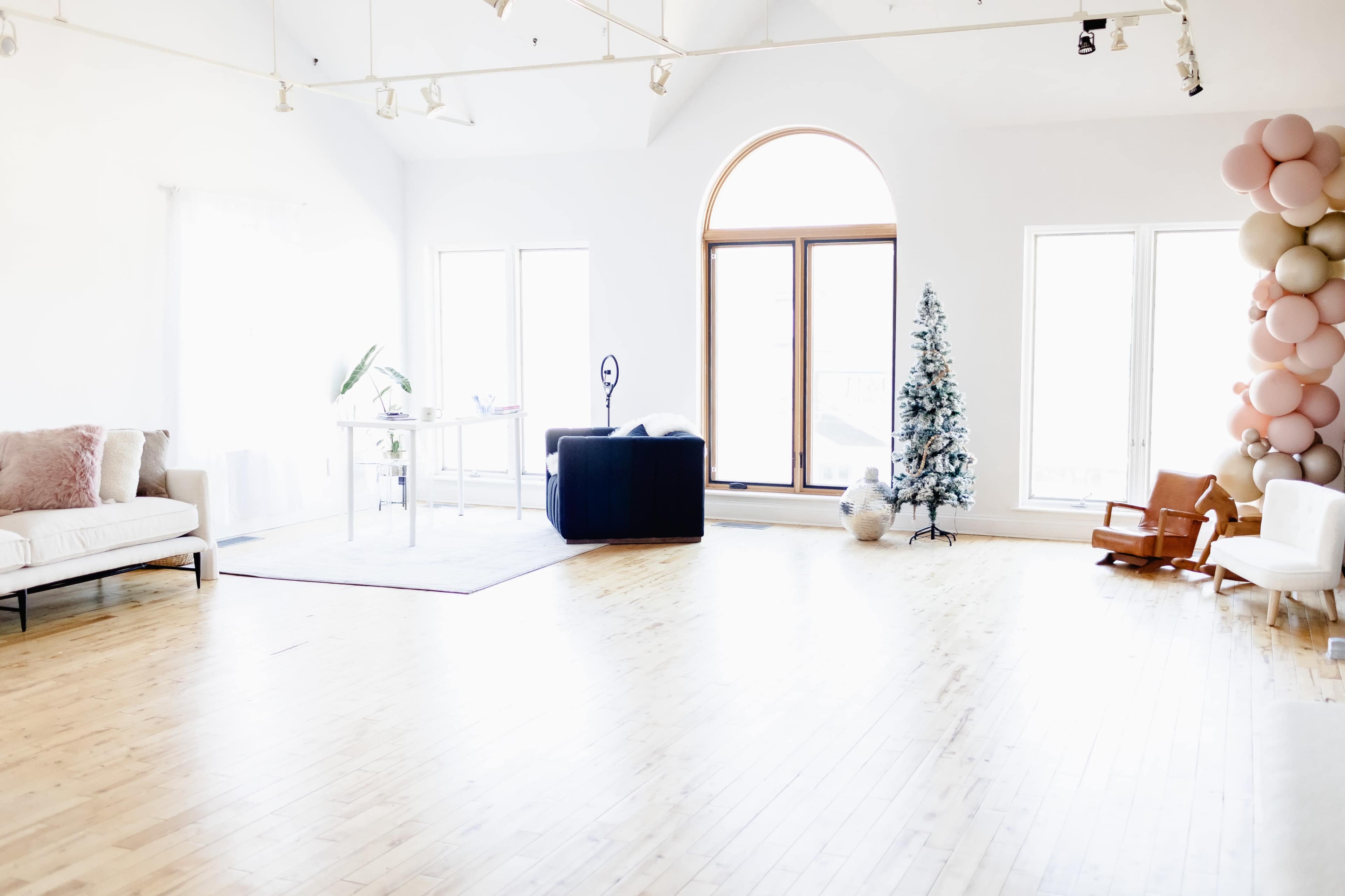 A spacious, well-lit room with wooden flooring, featuring a Christmas tree, a desk with a chair, and a seating area with a couch and a rocking chair.
