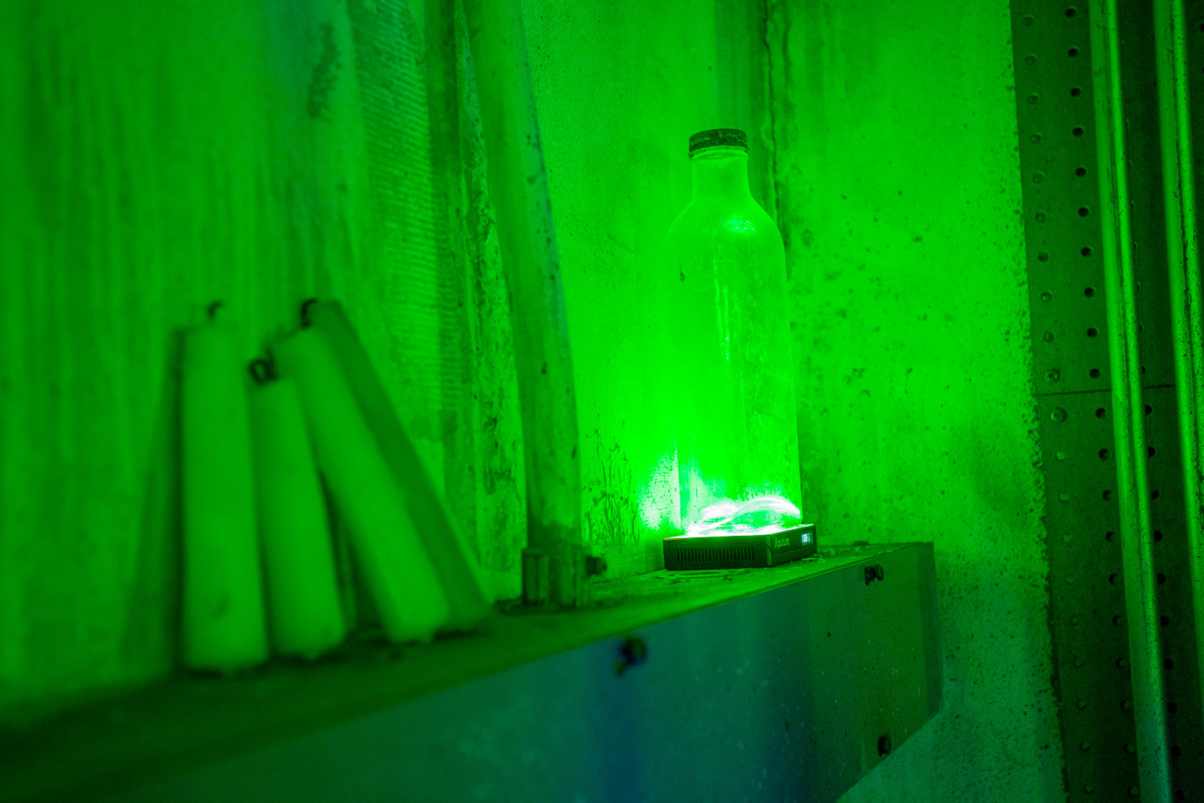 A glowing green bottle sits on a shelf next to several white objects in a dimly lit room.