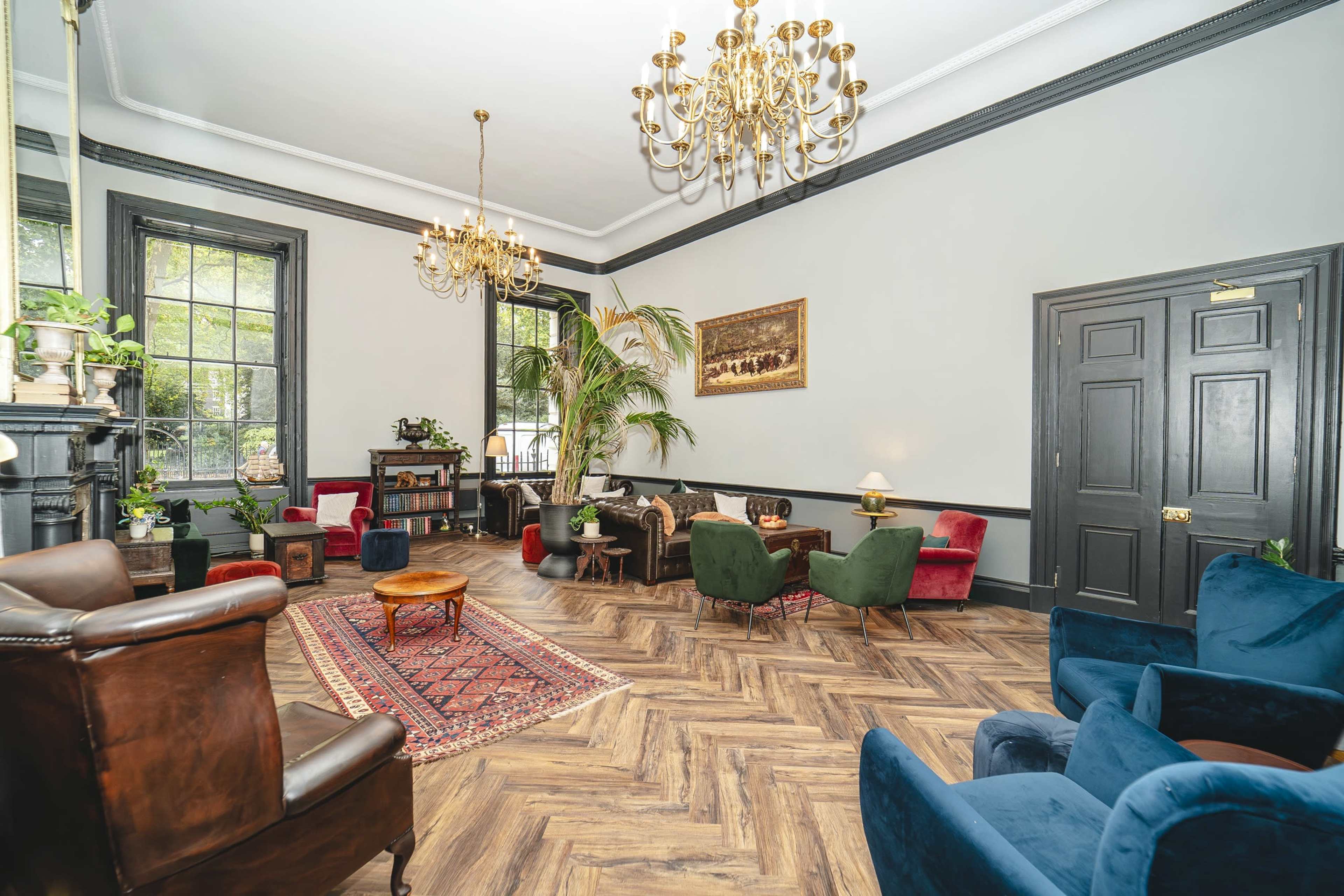 The interior of a stylish lounge features a mix of colorful seating, a chandelier, and decorative elements, with hardwood floors and large windows letting in natural light.