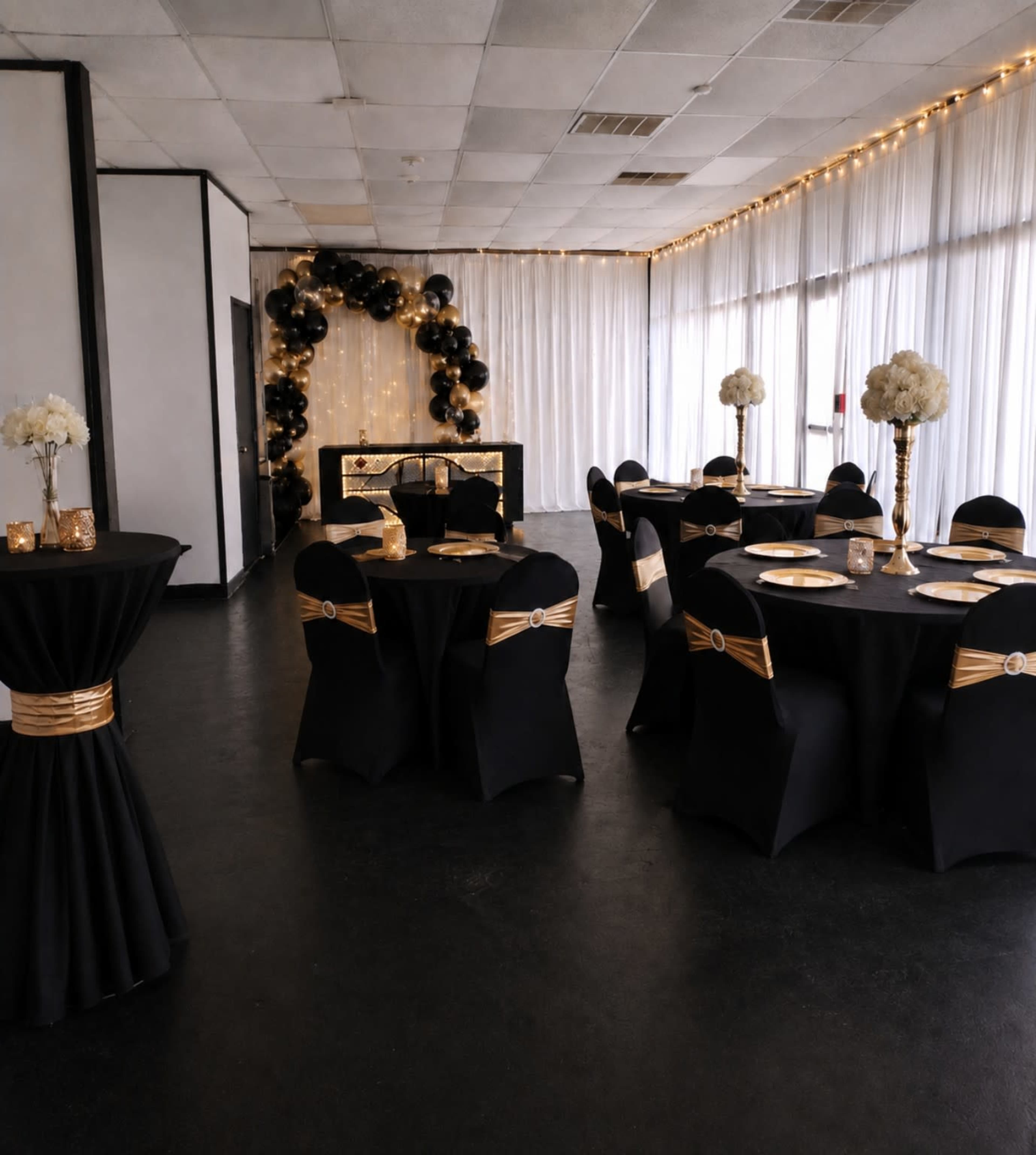 The image shows a decorated event space with black and gold table settings, featuring round tables, chairs adorned with gold sashes, and a backdrop of balloons.