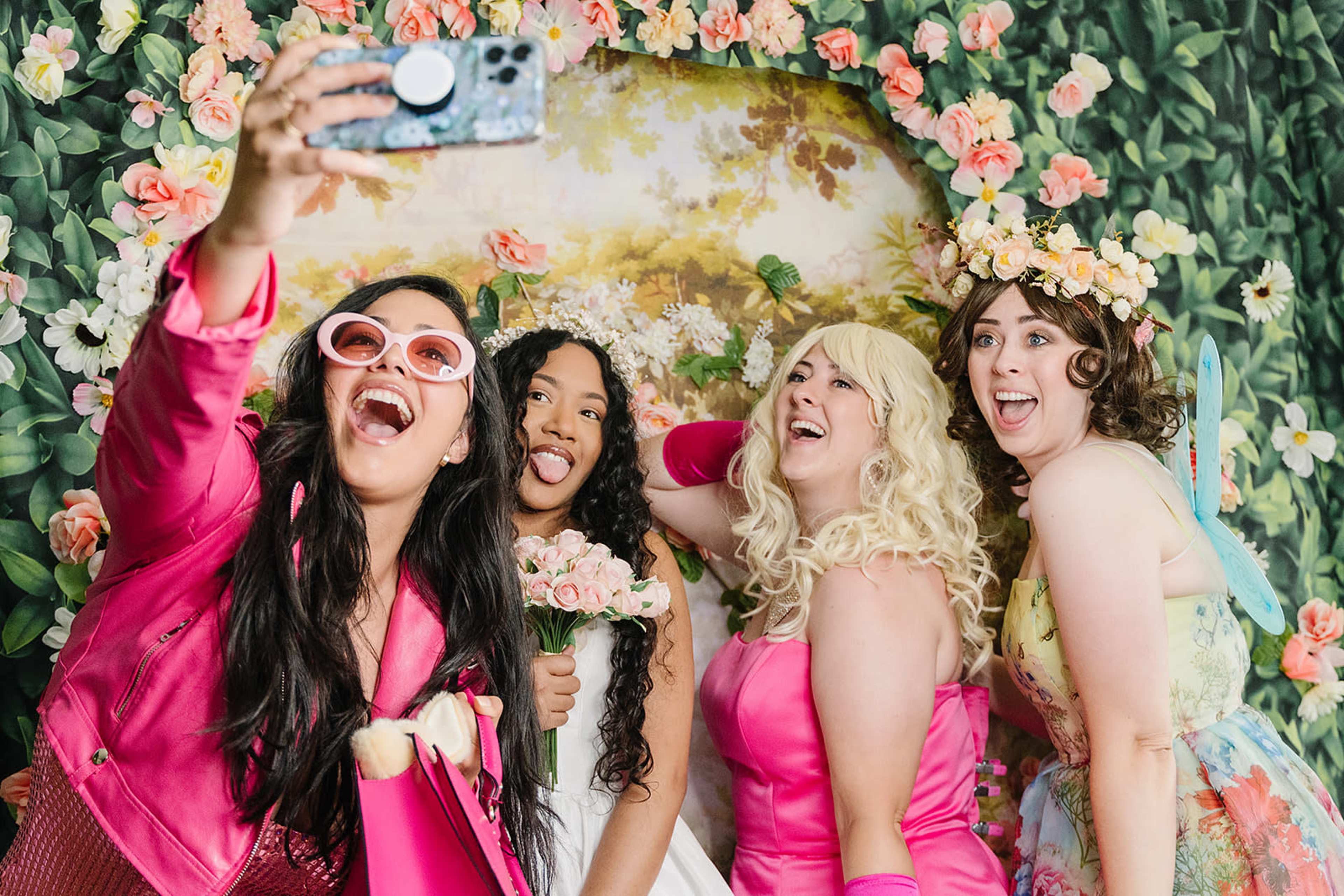 Four women pose for a selfie in front of a floral backdrop, with one holding a bouquet and another wearing a flower crown.