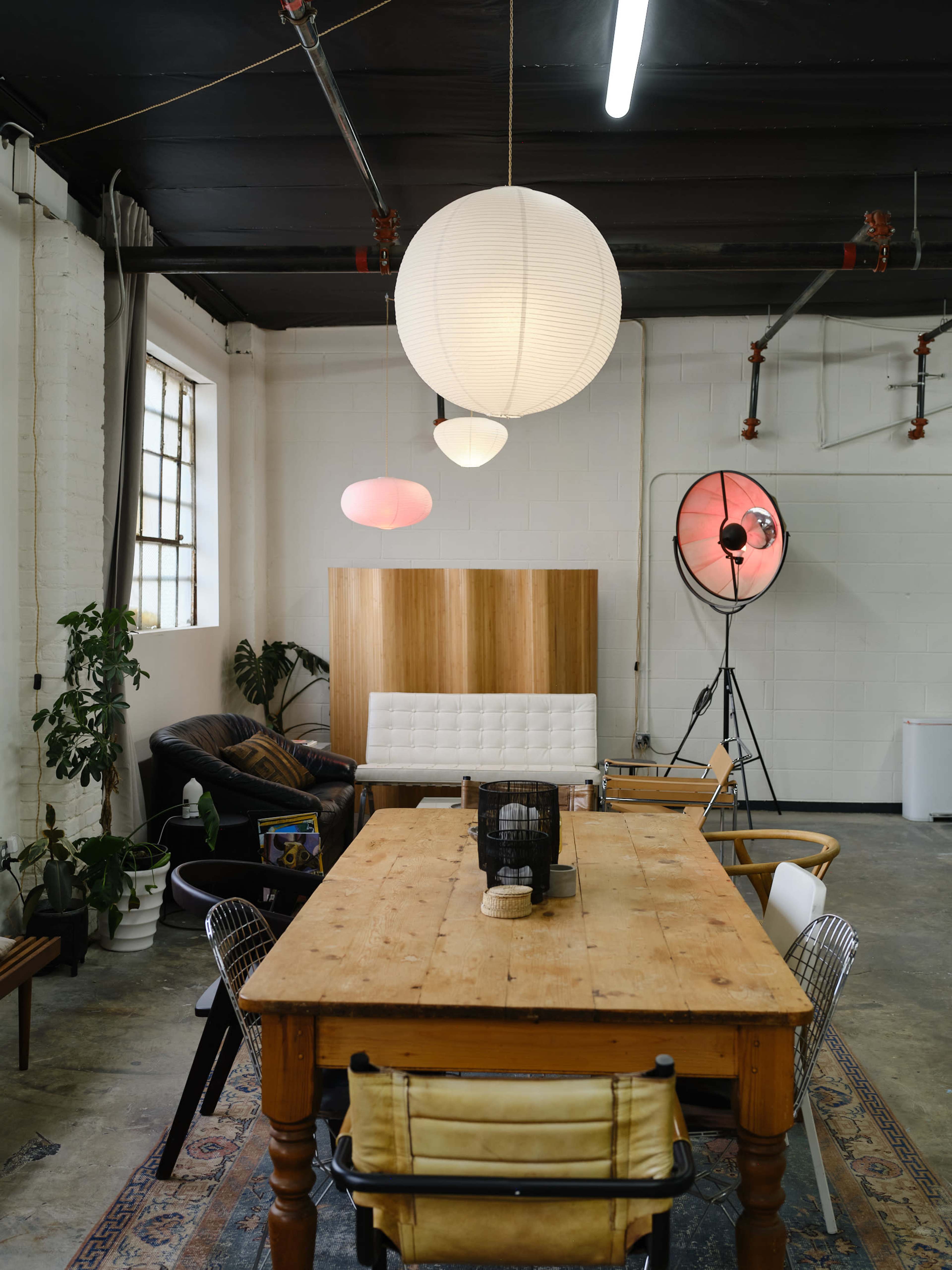 A spacious workspace features a wooden dining table surrounded by various chairs, illuminated by pendant lamps, with a cozy seating area and a large softbox light in the corner.