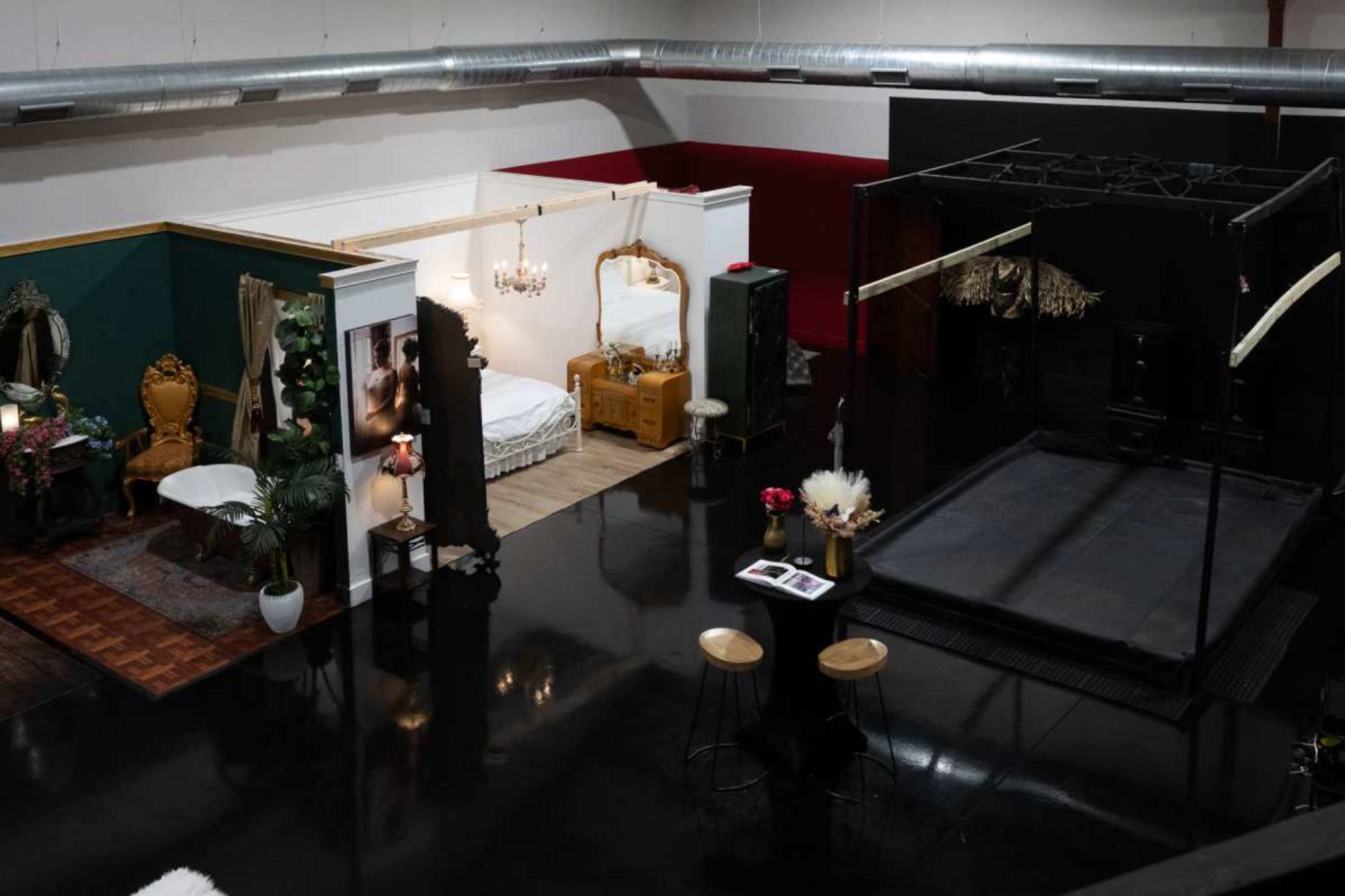 The image shows a spacious indoor studio featuring distinct areas for photography, including a bedroom setup with a bed and decor, a vintage-inspired sitting area, and a black stage with a raised platform.