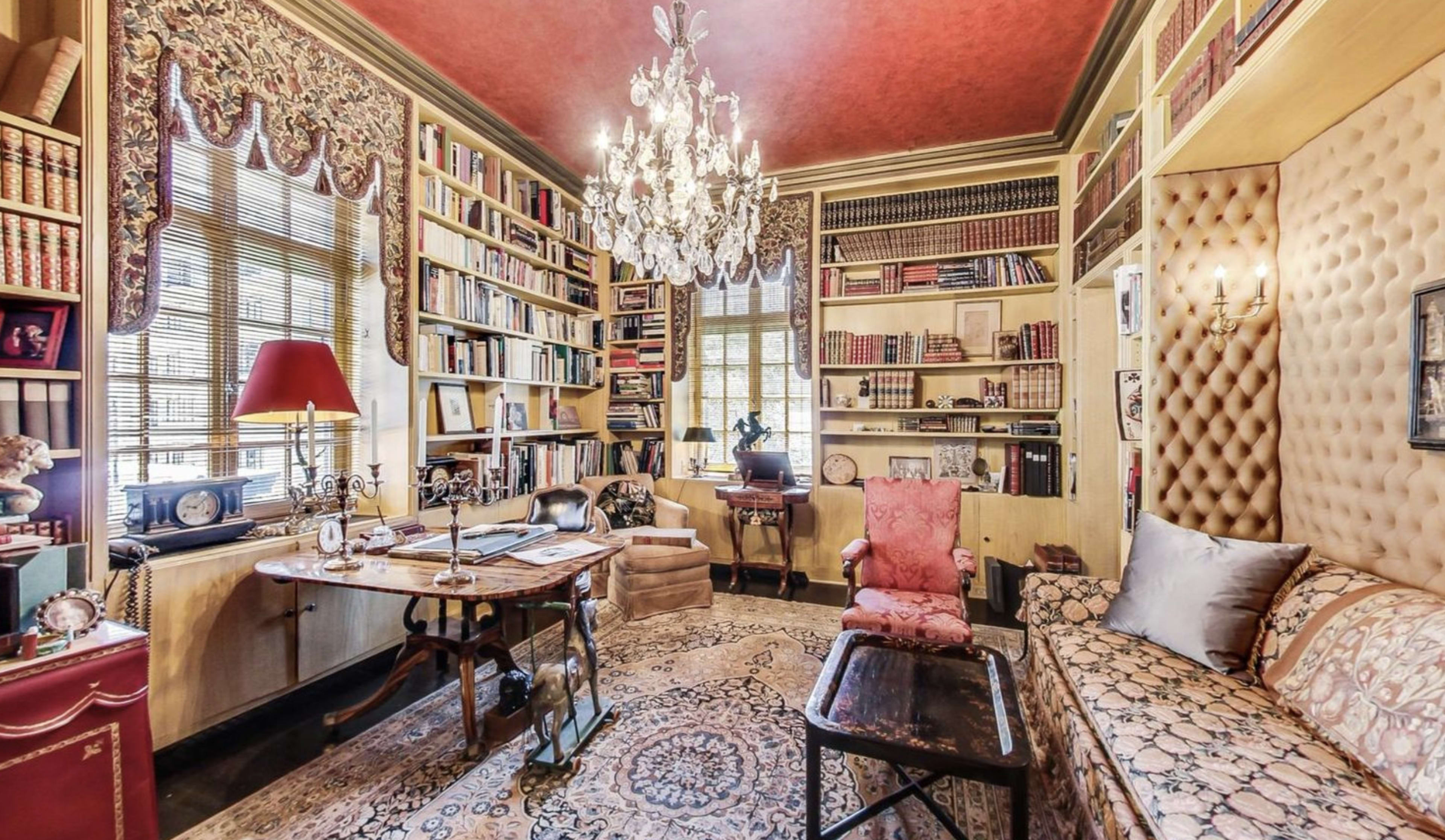 The image shows a cozy home office filled with bookshelves, a large desk, and a chandelier, featuring a decorative rug and comfortable seating.