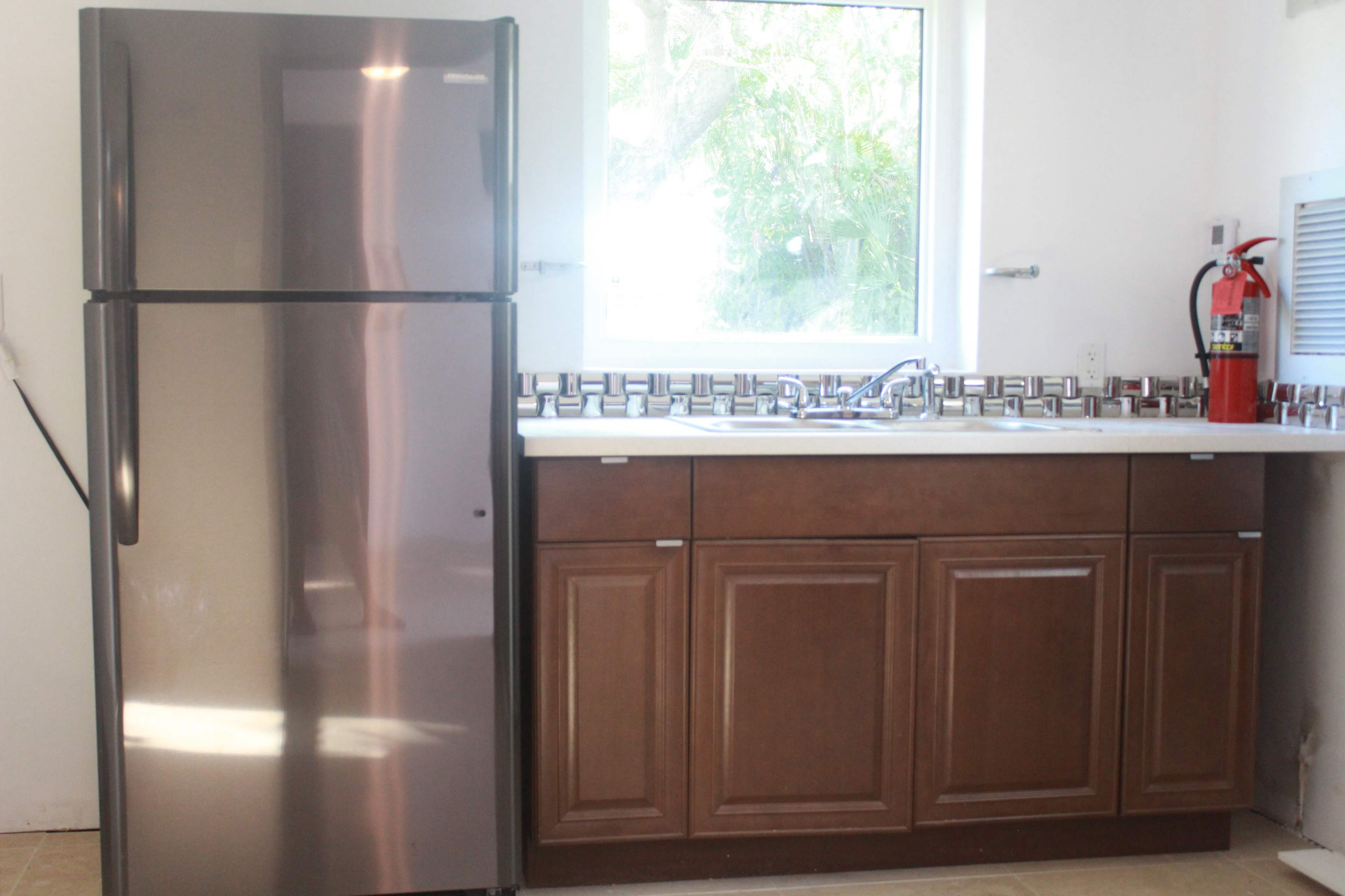 The image shows a kitchen area featuring a stainless steel refrigerator next to a wooden cabinet with a sink and a fire extinguisher mounted on the wall.