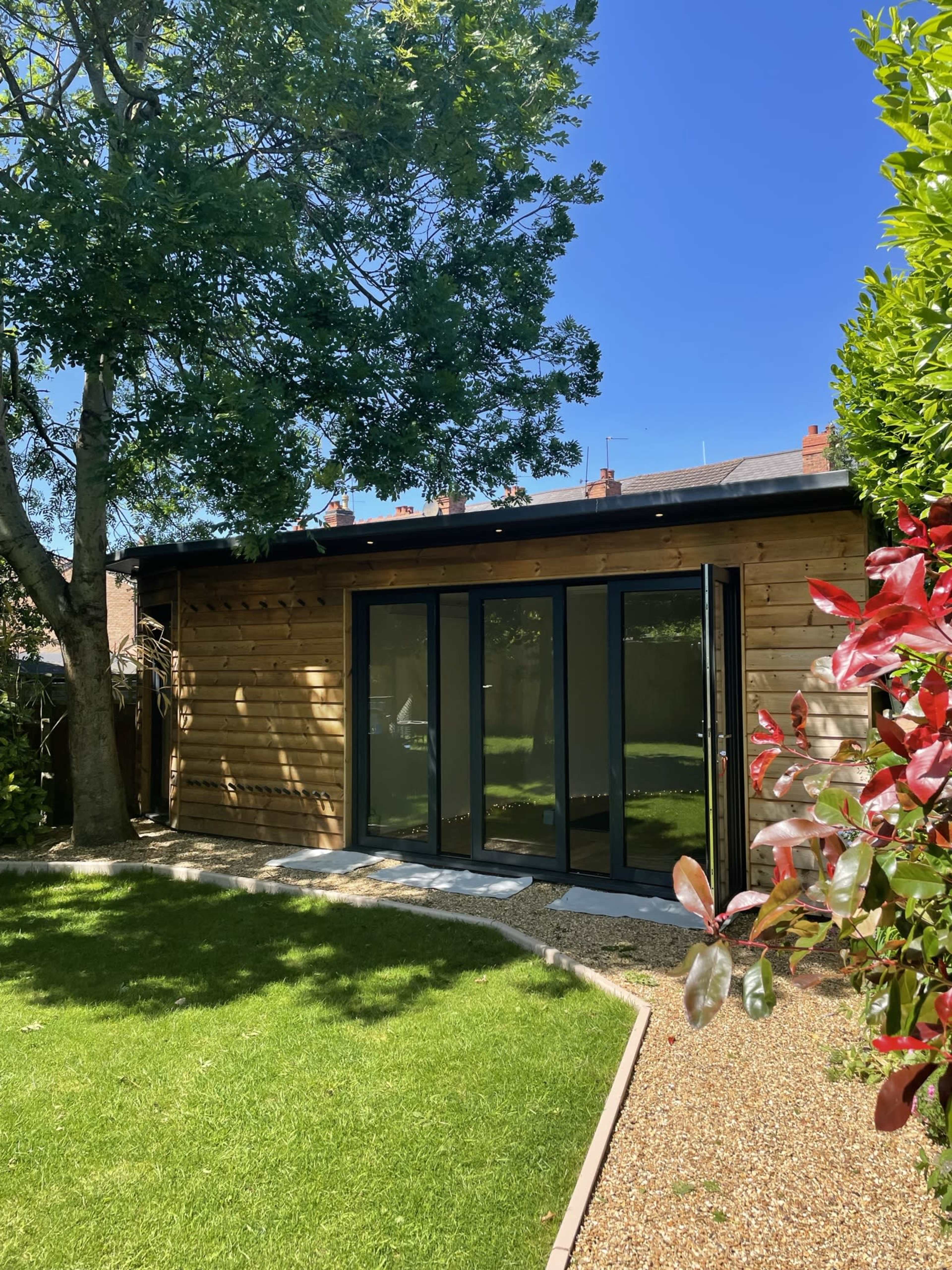A wooden garden shed with large glass doors is situated in a green backyard surrounded by trees and shrubbery.