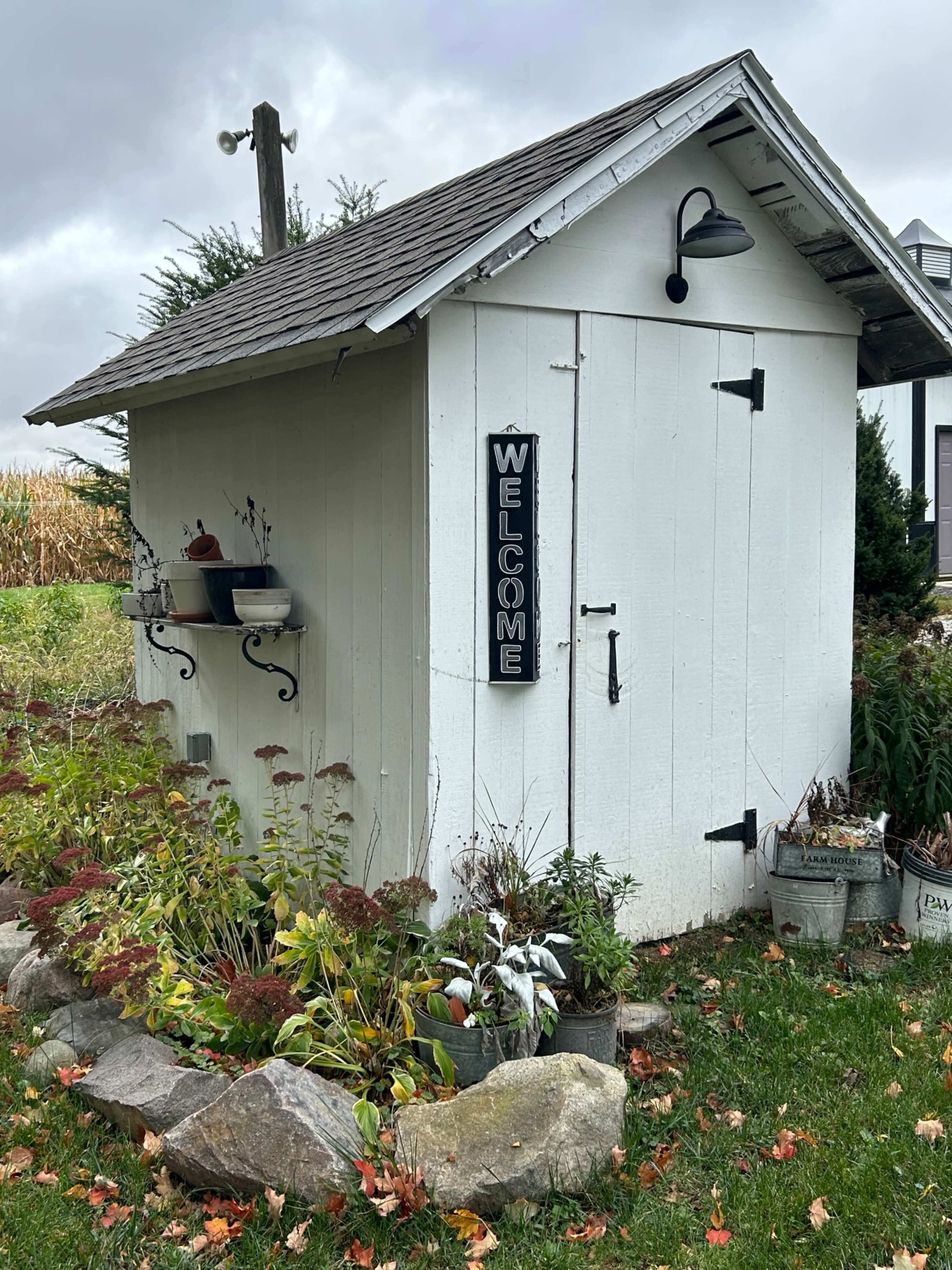A small white shed with a "WELCOME" sign is surrounded by colorful plants and rocks in a garden setting.
