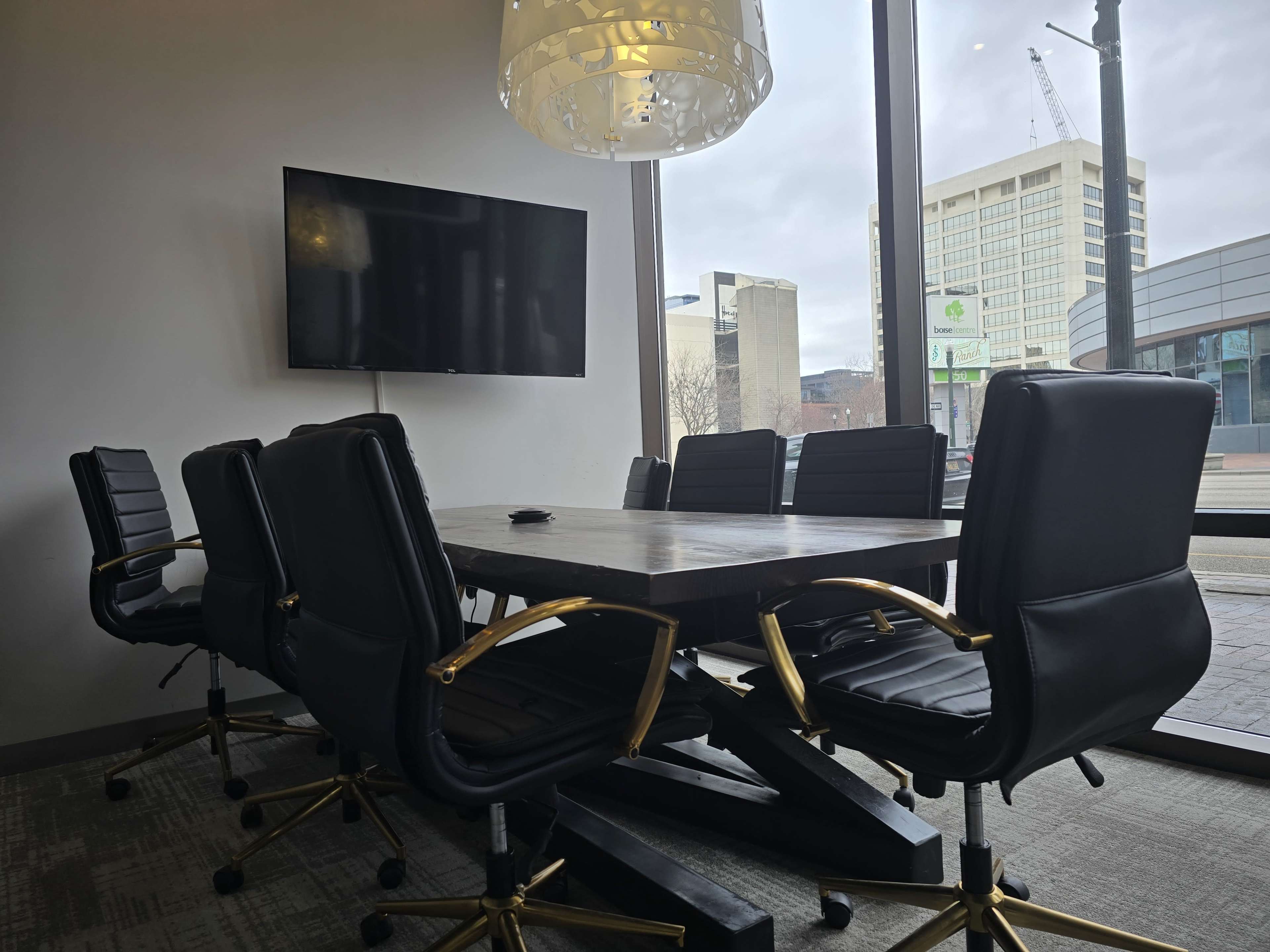 A modern conference room features a wooden table surrounded by black rolling chairs, with a large TV screen mounted on the wall and windows providing a view of the city.