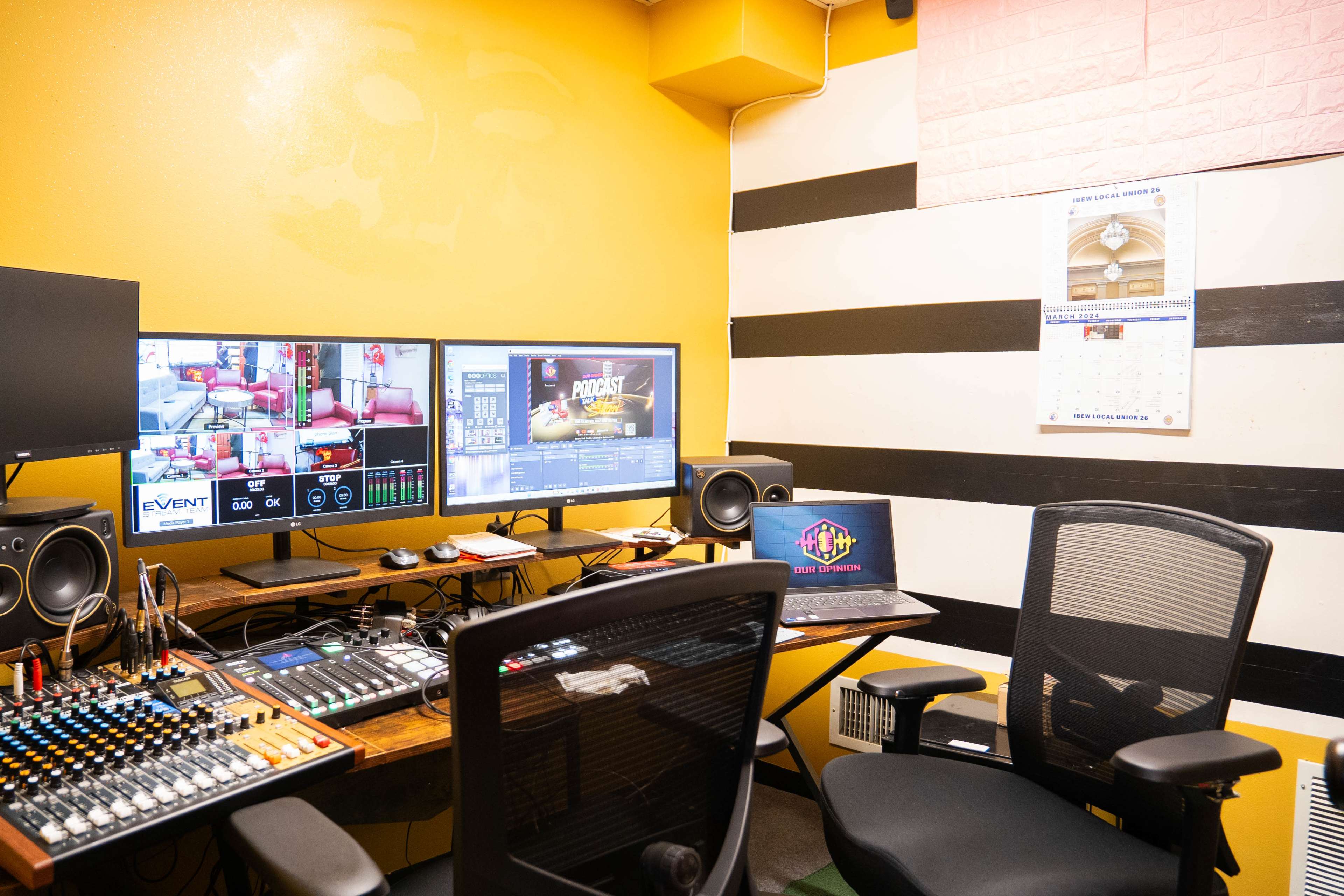 A studio setup with two large monitors, a mixing console, a laptop, and yellow-painted walls, alongside a calendar on the wall.