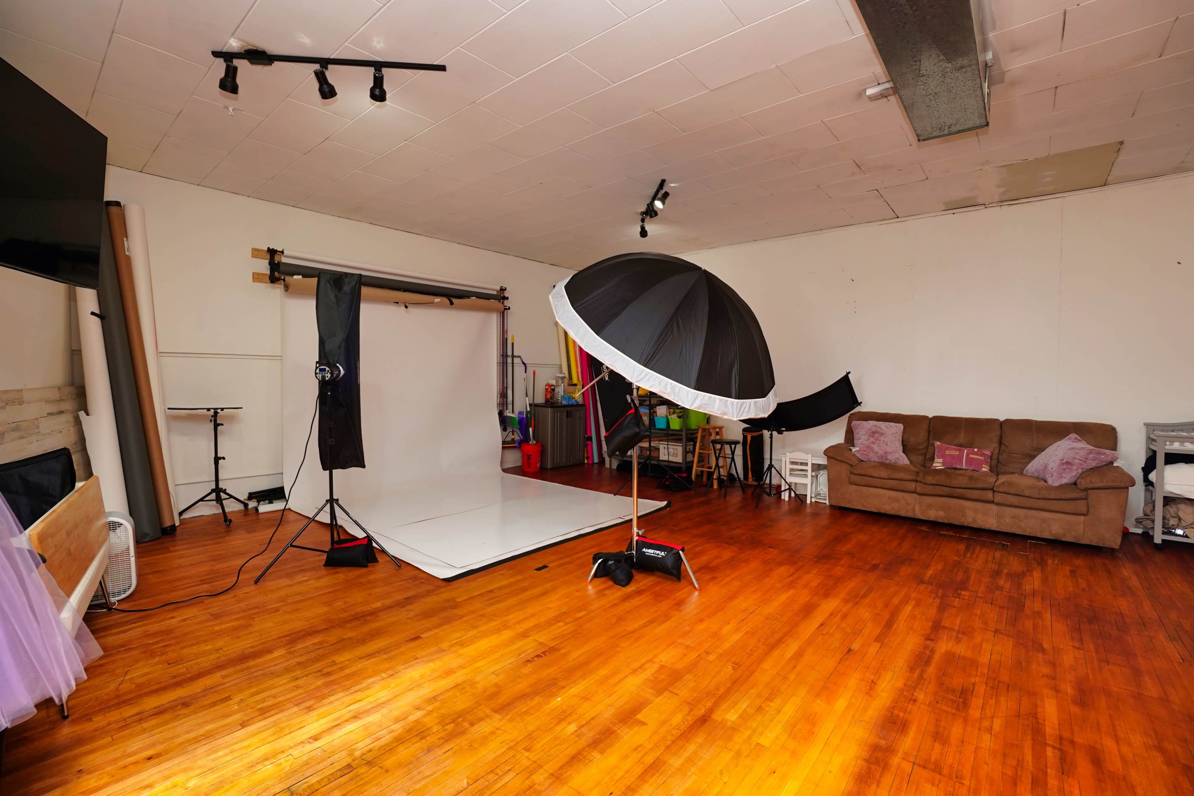 A photography studio with a white backdrop, an umbrella light setup, and a brown couch against the wall.