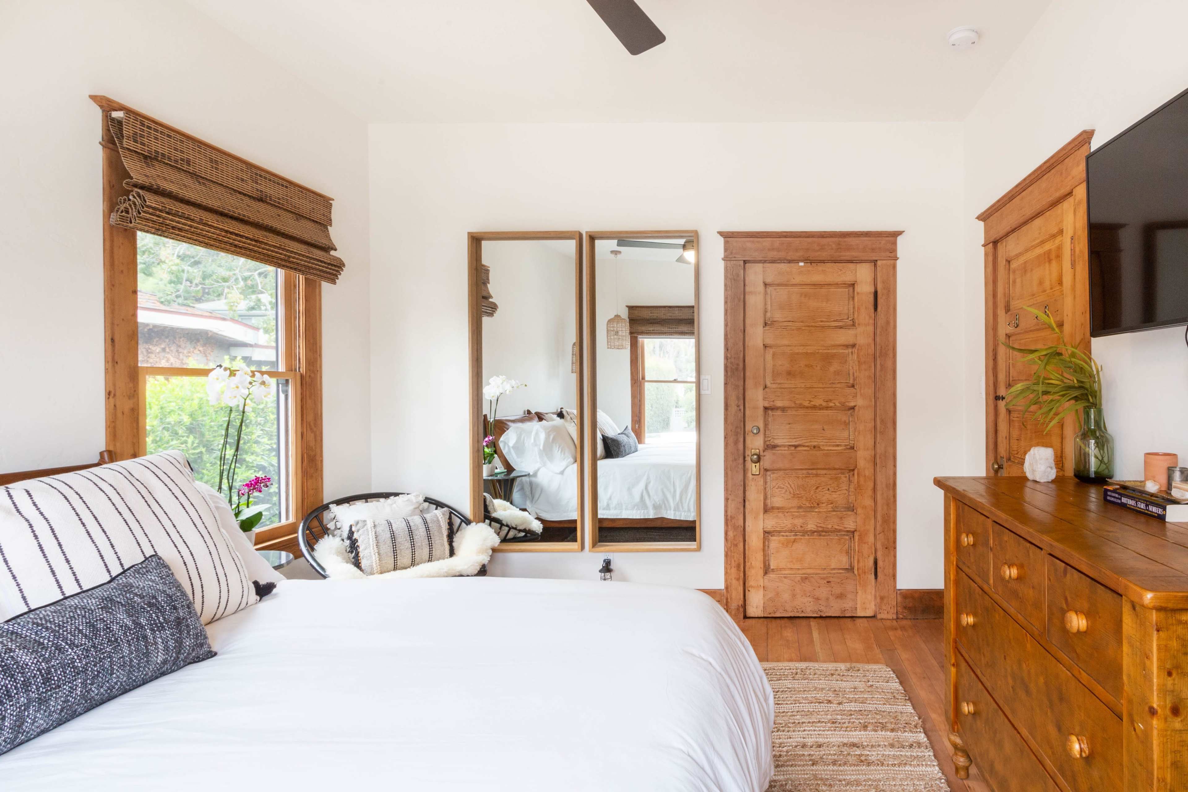 A bright bedroom featuring a bed, a wooden dresser, a large mirror, and a window with woven shades.