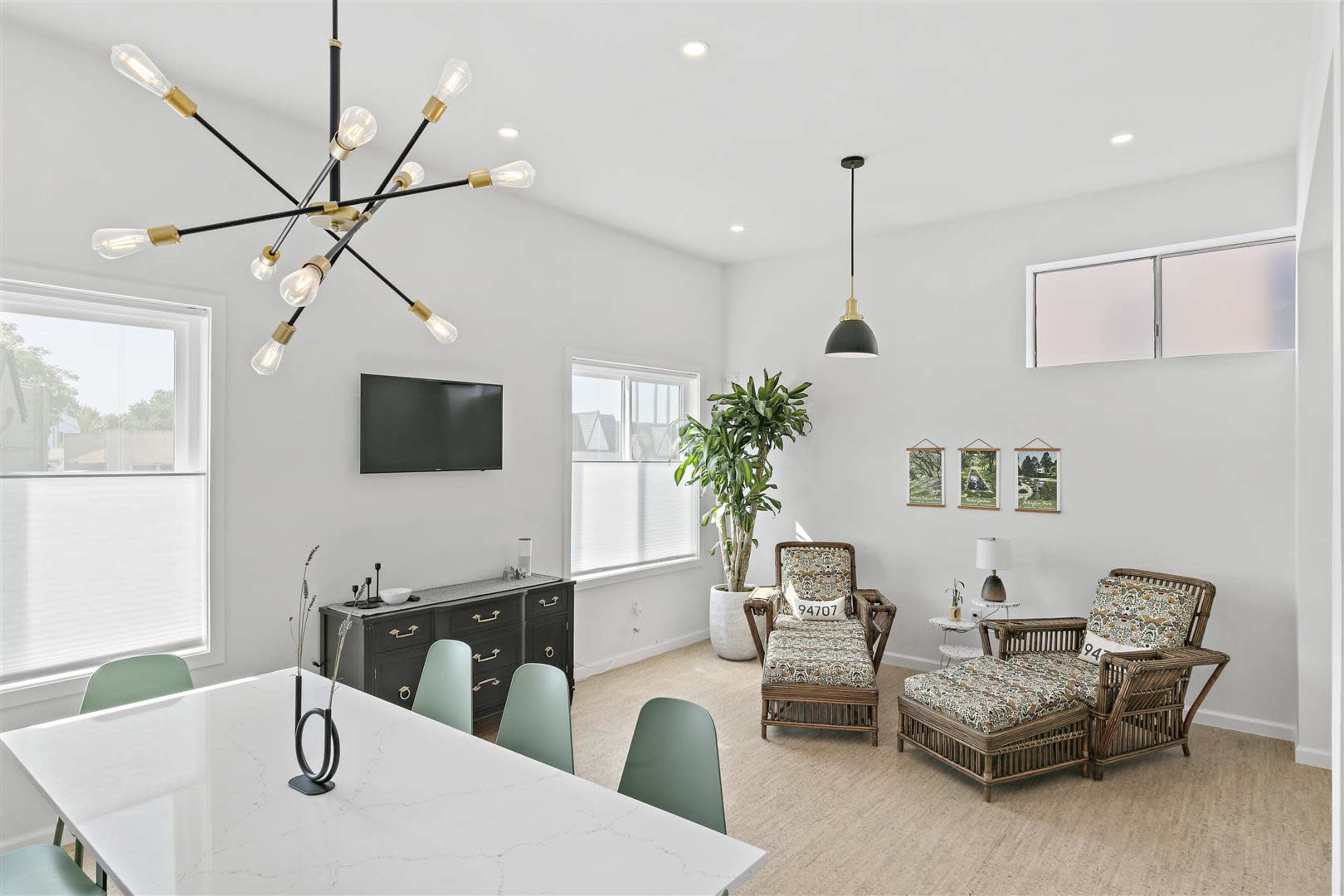 A bright living area features a modern dining table, a television mounted on the wall, and comfortable seating with a large window and a potted plant.
