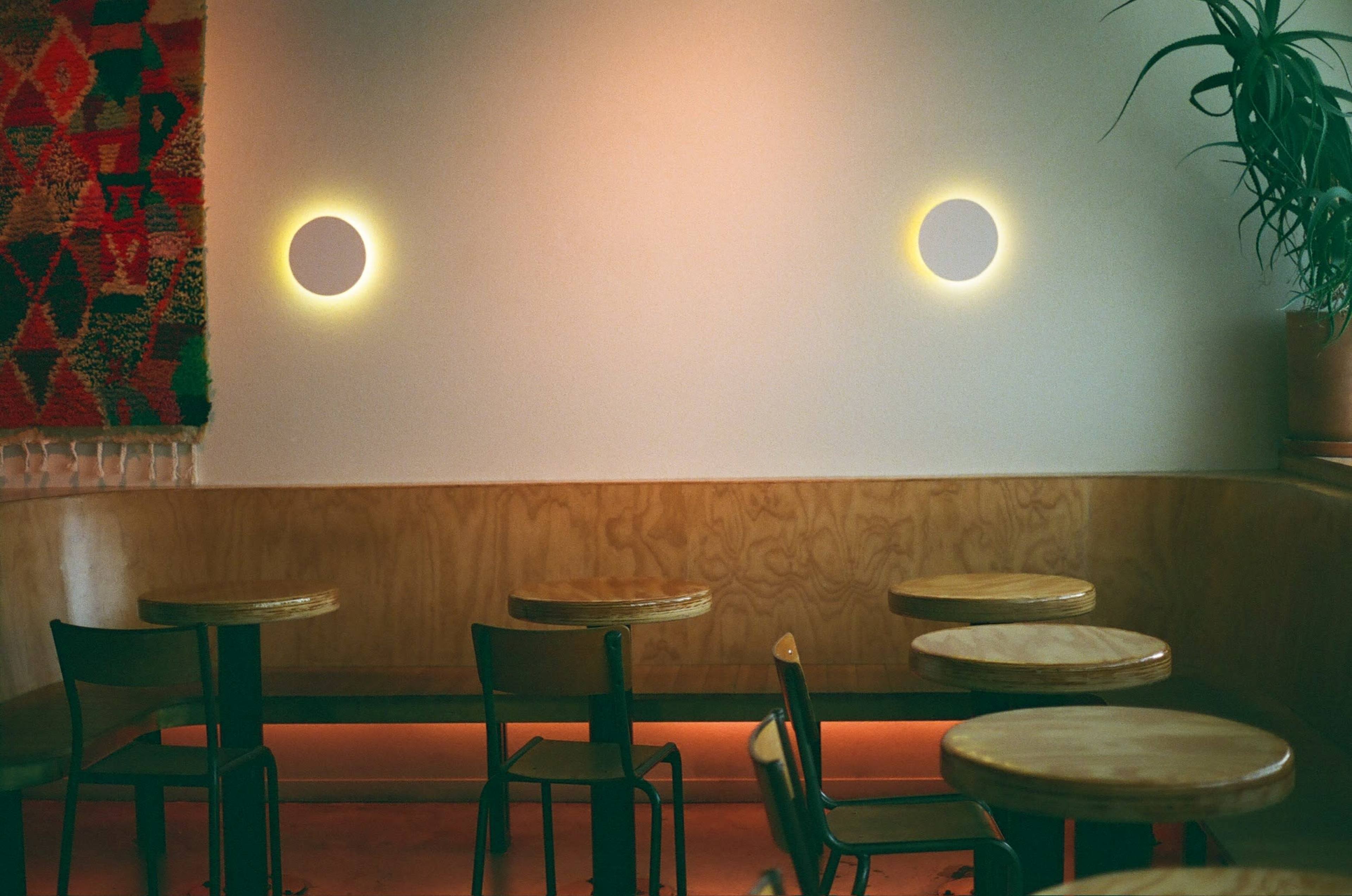 The image shows a minimalist cafe interior with wooden tables and chairs, accented by two circular wall lights and a textured tapestry on the wall.