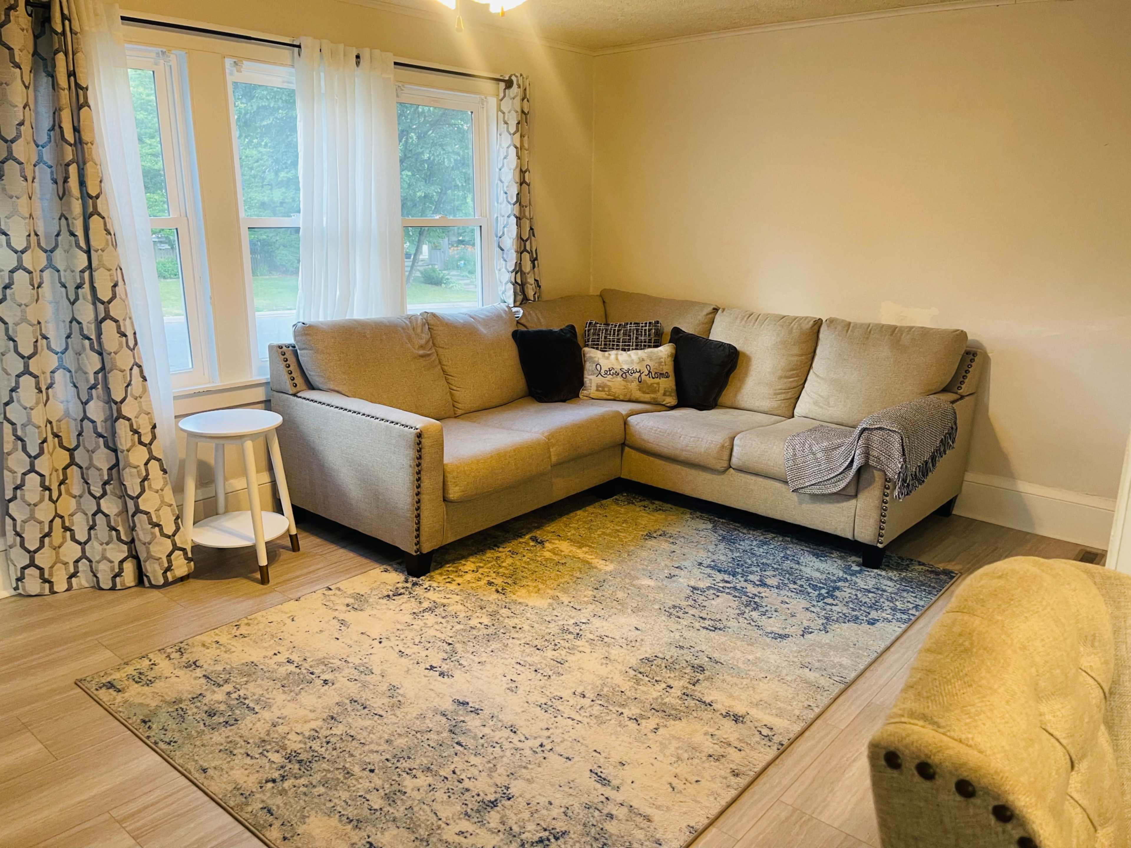 A cozy living room with a sectional sofa, a round side table, and a large area rug, all positioned beside two windows with sheer curtains.