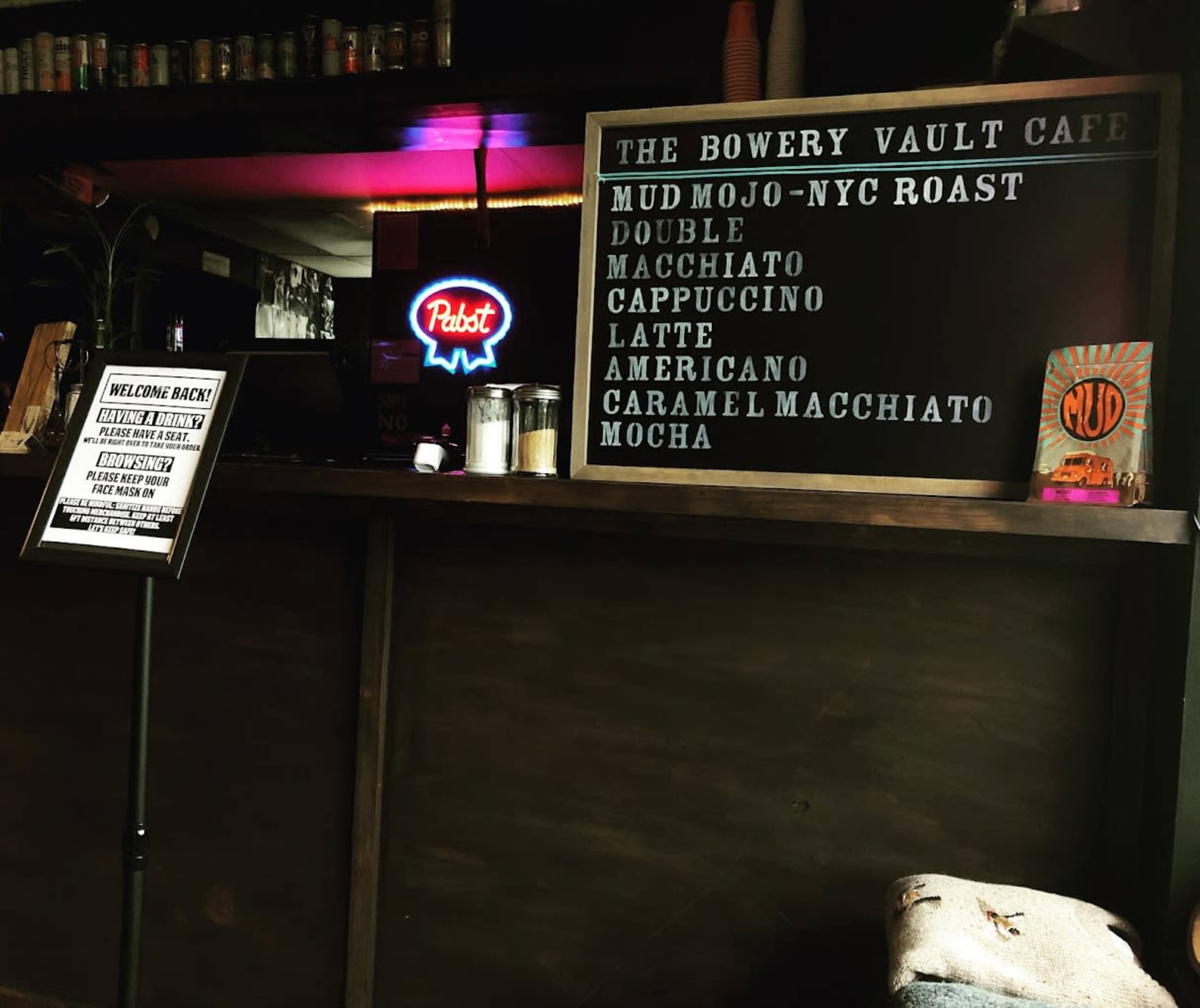 The image features a coffee shop counter with a menu board displaying various coffee options, alongside a neon sign and a can of beer.