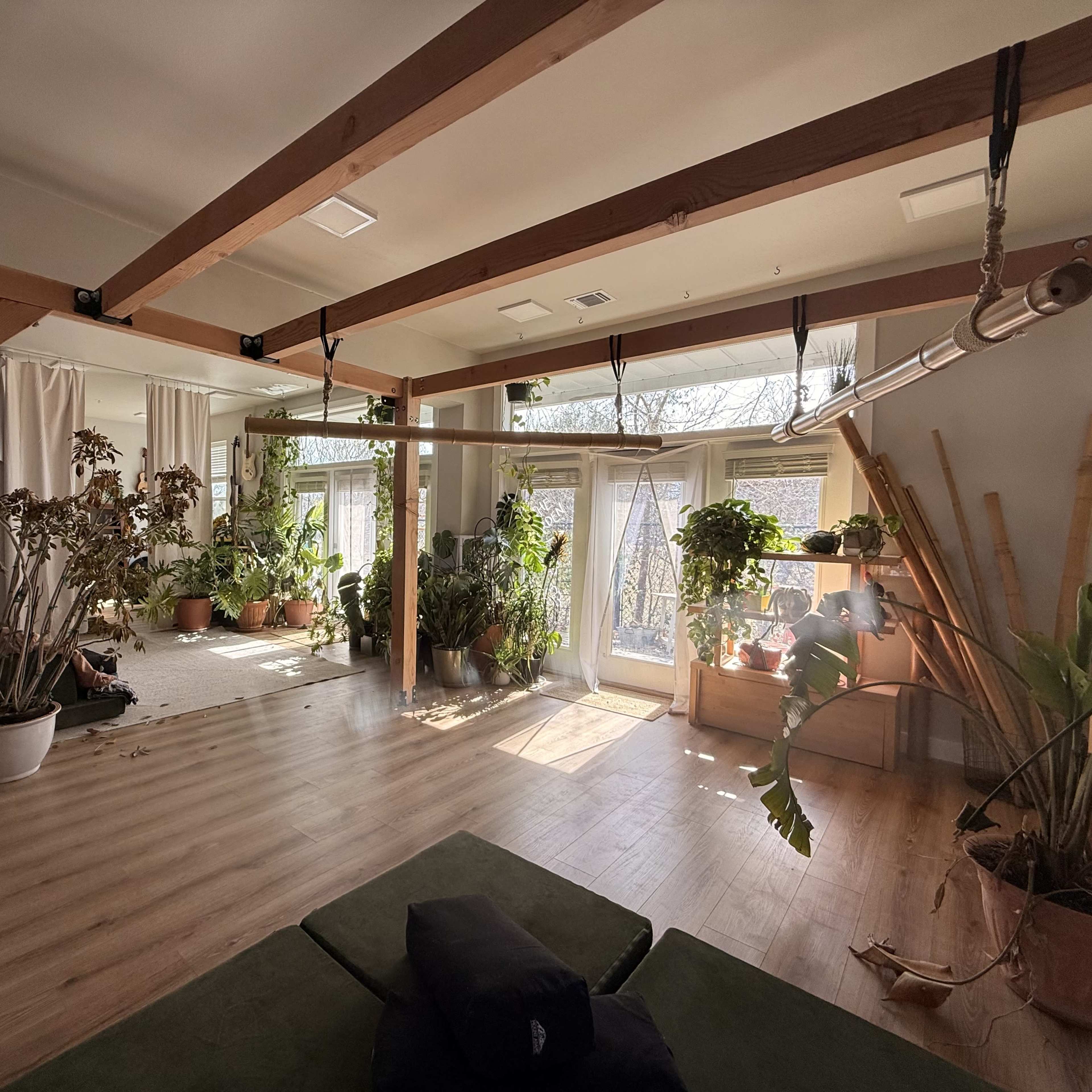 A bright and spacious indoor room features large windows, green plants in various pots, and wooden beams supporting the ceiling.