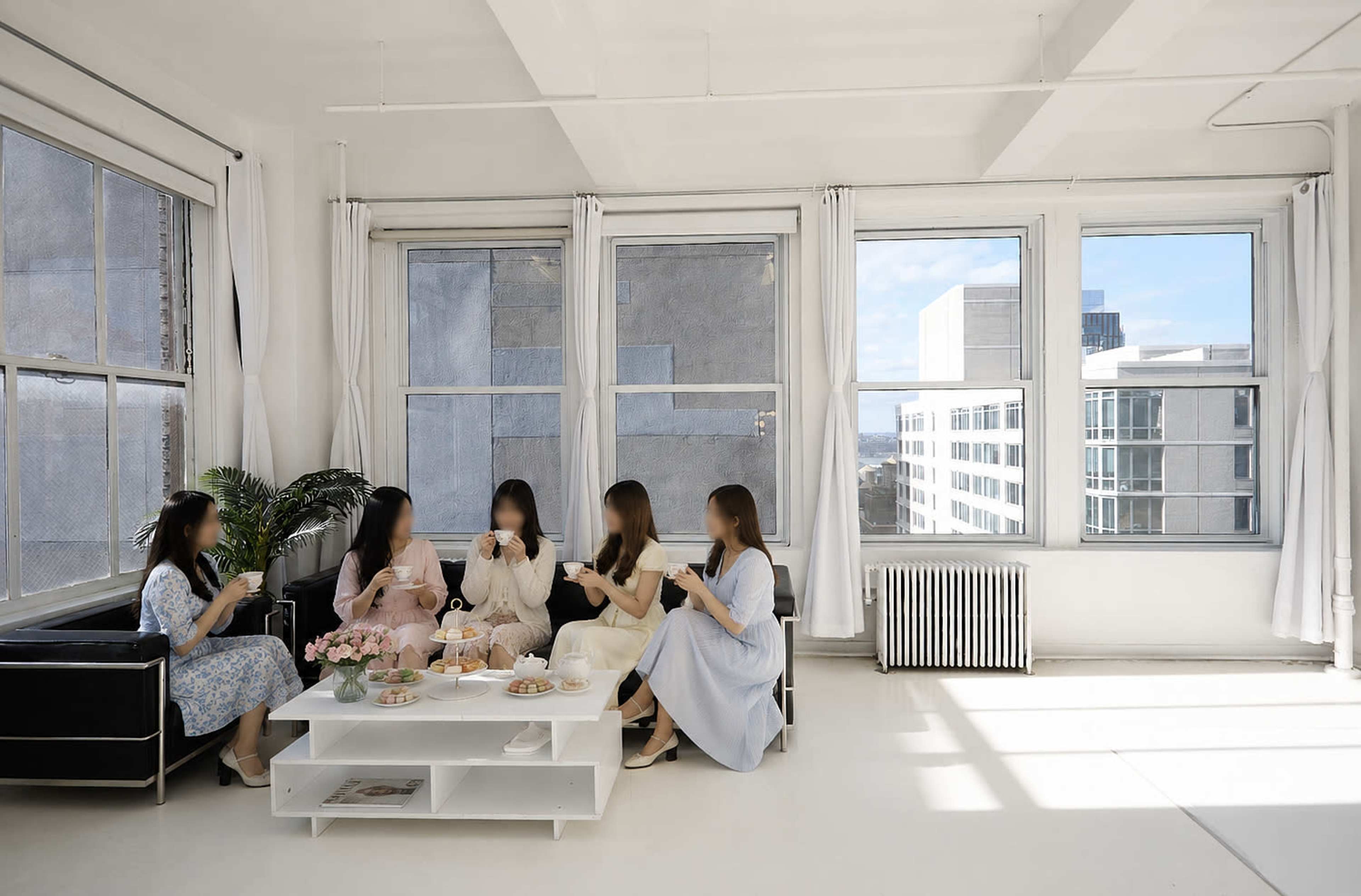 Five women are seated around a white coffee table in a bright, airy room with large windows and a view of a modern building outside.