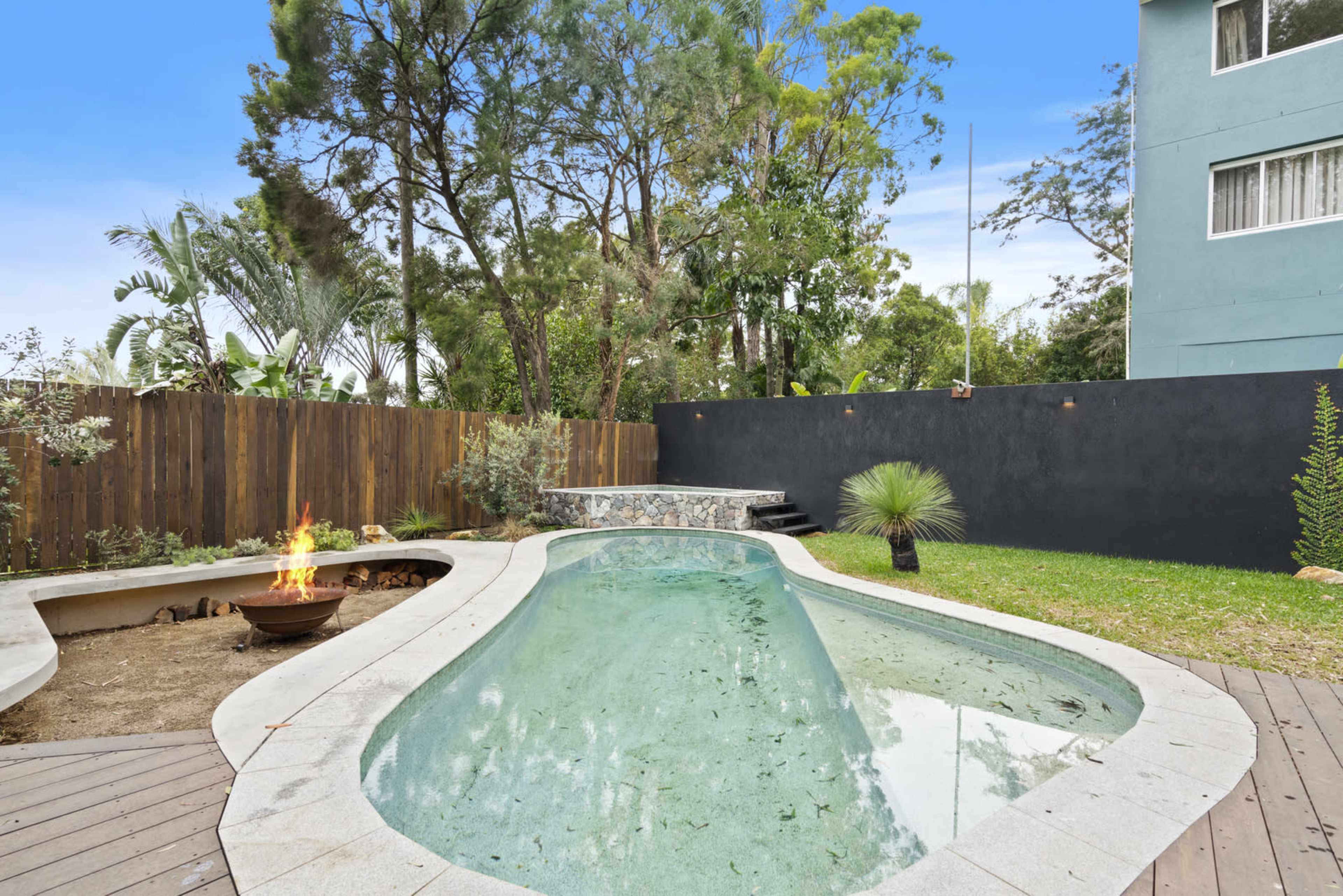 The image shows a landscaped backyard featuring a pool surrounded by trees, a fire pit, and a dark wall, with a two-story building in the background.