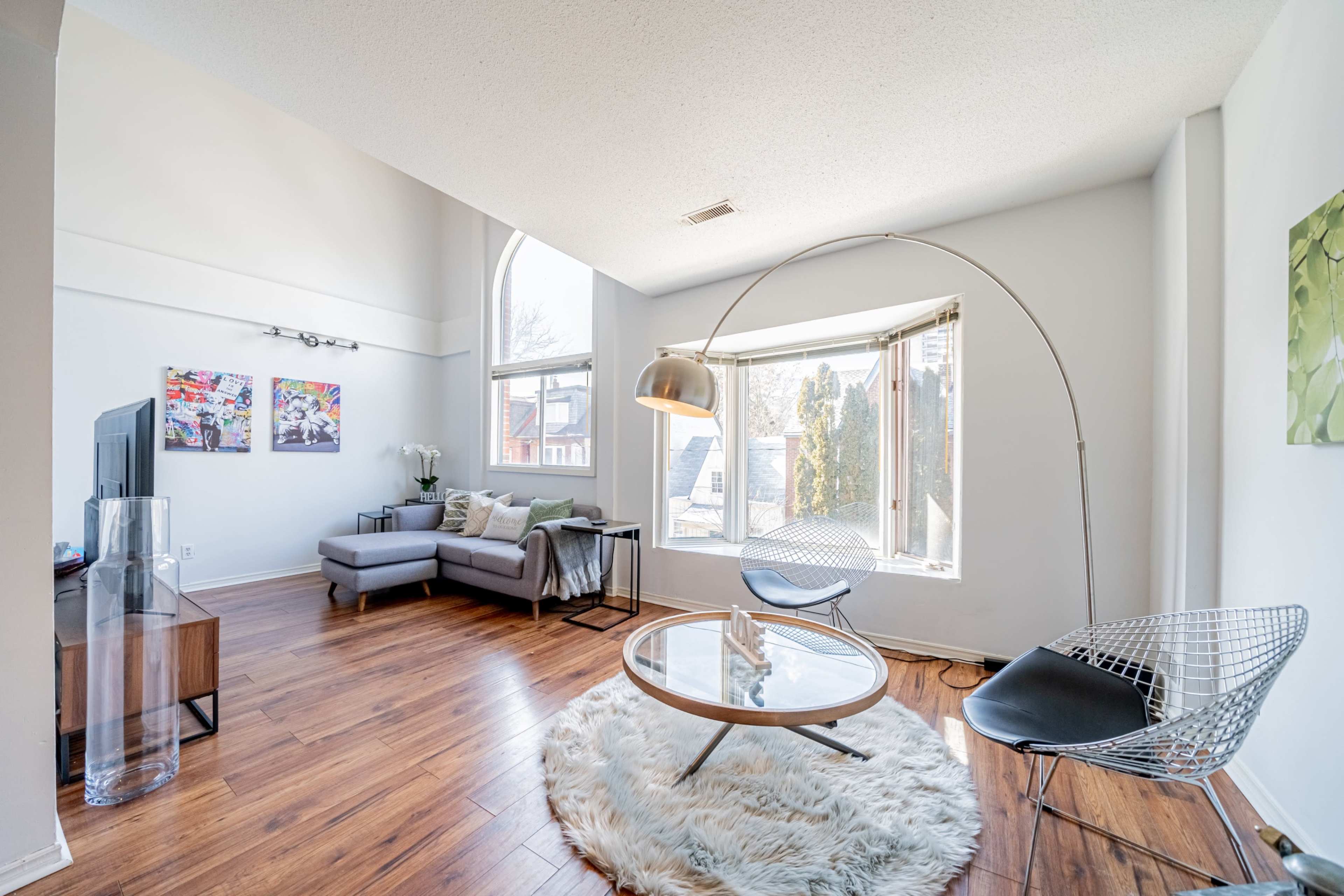 A modern living room with a gray sofa, a circular glass coffee table, a large floor lamp, and two distinct seating areas near a bay window.