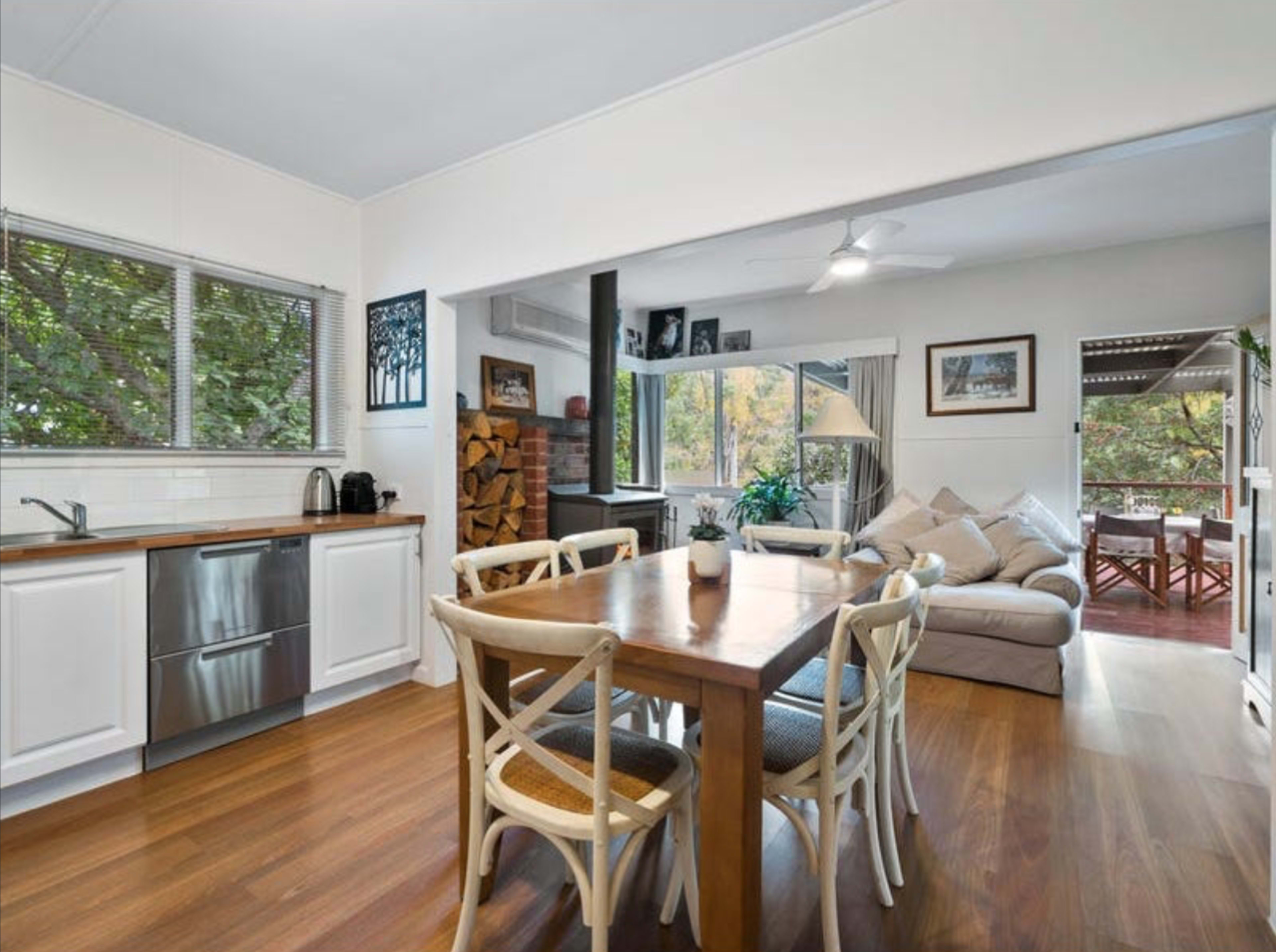 A dining area with a wooden table and chairs opens to a living space featuring a couch and stacked firewood in the background.