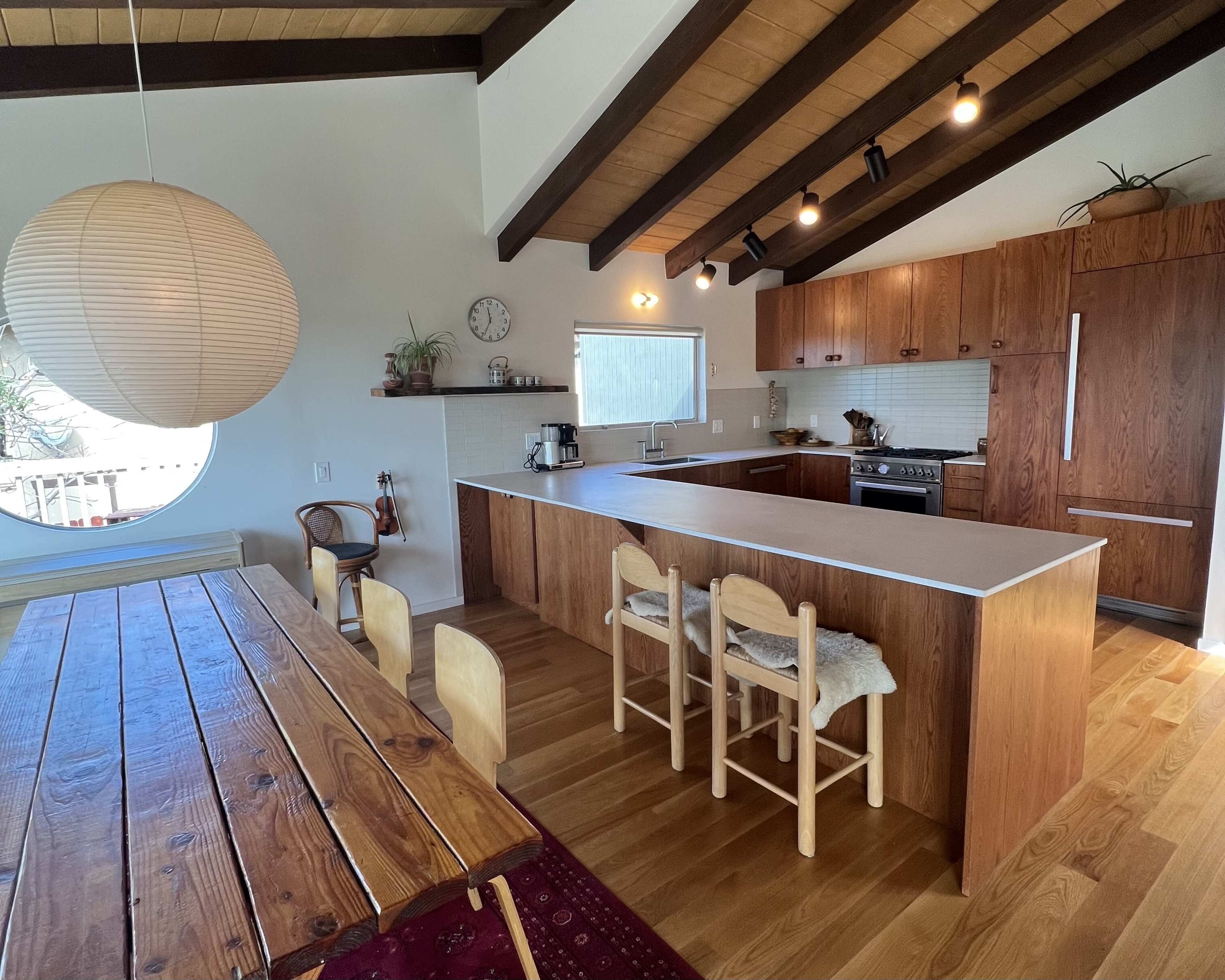The image shows a modern kitchen with wooden cabinetry, a large island with seating, and a dining table adjacent to the space.
