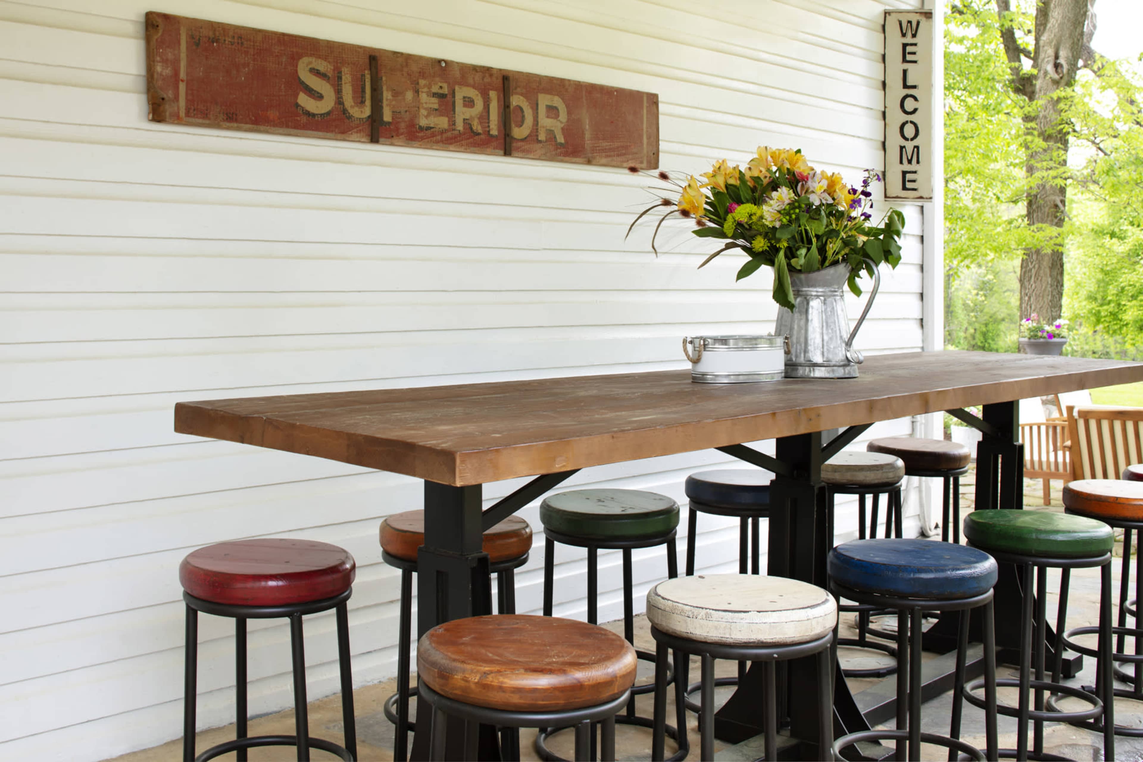 A rustic outdoor bar with a wooden countertop and multicolored stools is set against a white wooden wall, adorned with a welcome sign and a vase of flowers.