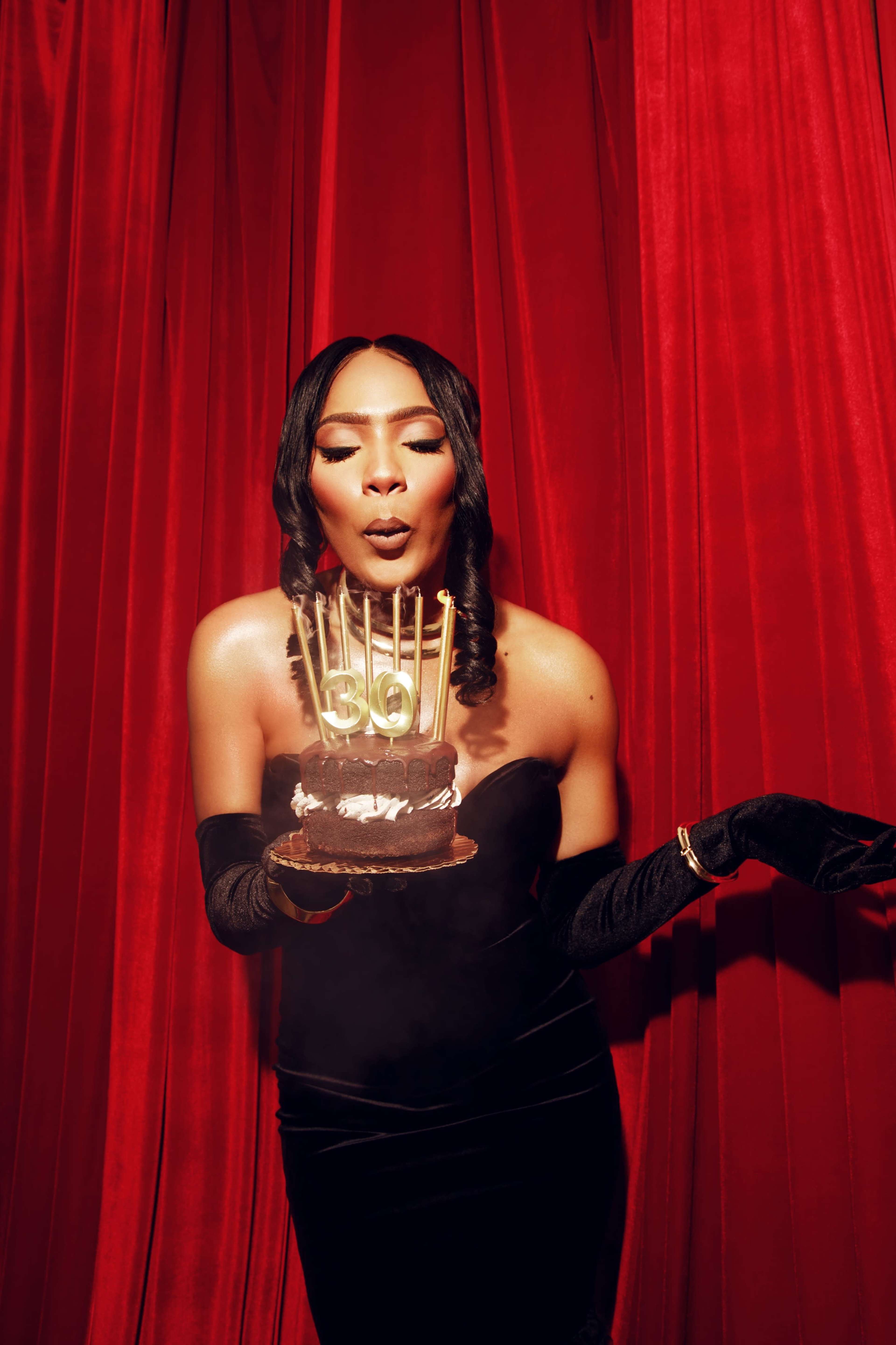 A person in a black dress holds a cake with candles against a red curtain backdrop.