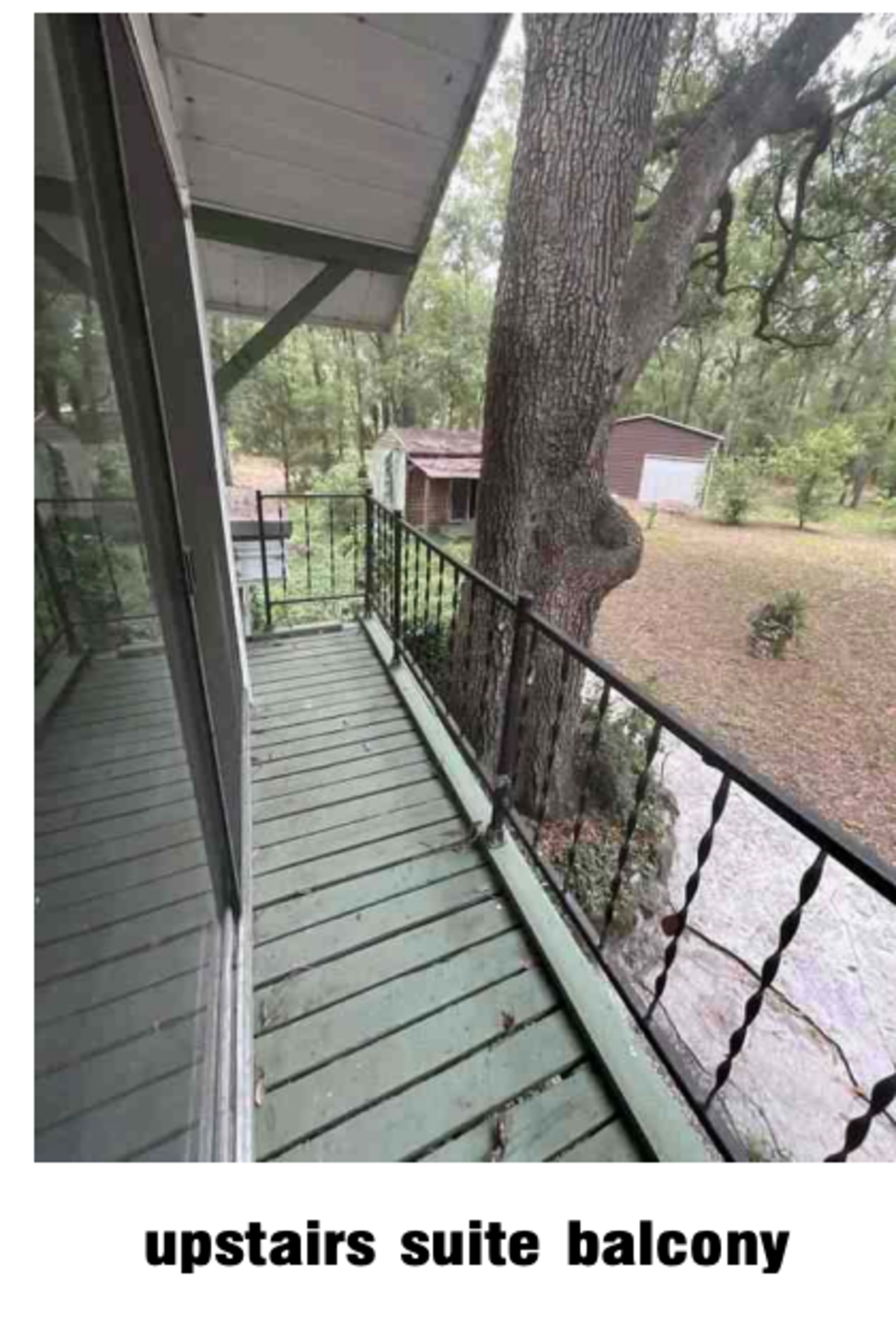 The image shows an upstairs balcony with a wooden floor and a wrought iron railing, overlooking a grassy area with trees and outbuildings.