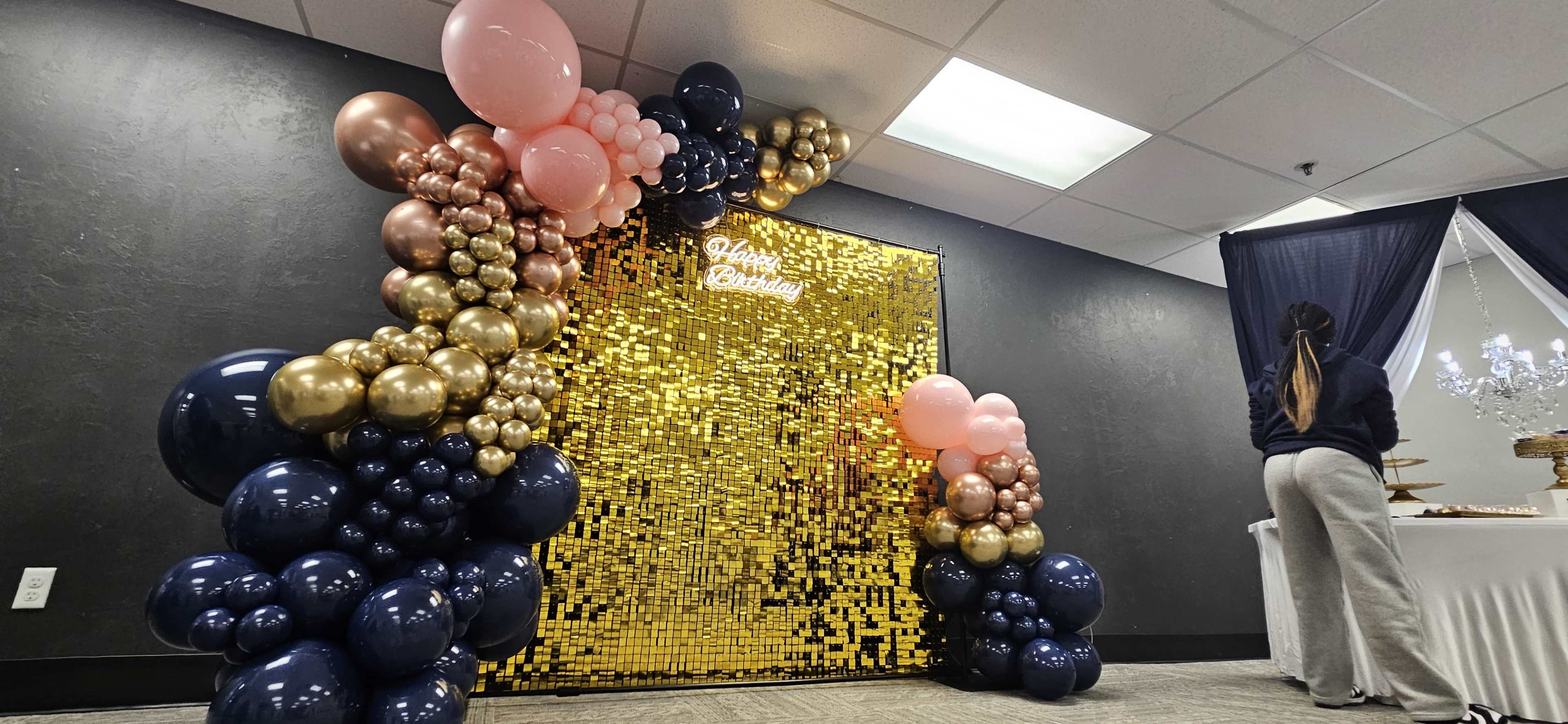 The image shows a decorative backdrop made of gold sequins surrounded by a balloon arch in pink, gold, and blue hues, with a person in gray pants standing beside a table set with food.